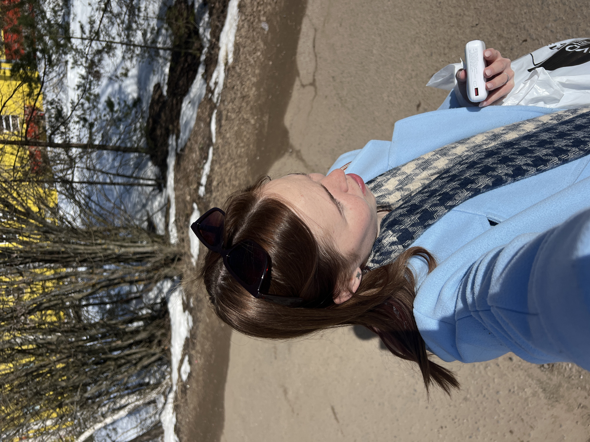 A woman with brown hair and sunglasses on her head is facing upwards, holding a white power bank in her right hand, with a white plastic bag nearby. She is wearing a light blue coat and a patterned scarf. The background shows a path with patches of snow, trees, and a yellow building. The location is Chaikovsky, Russia.
