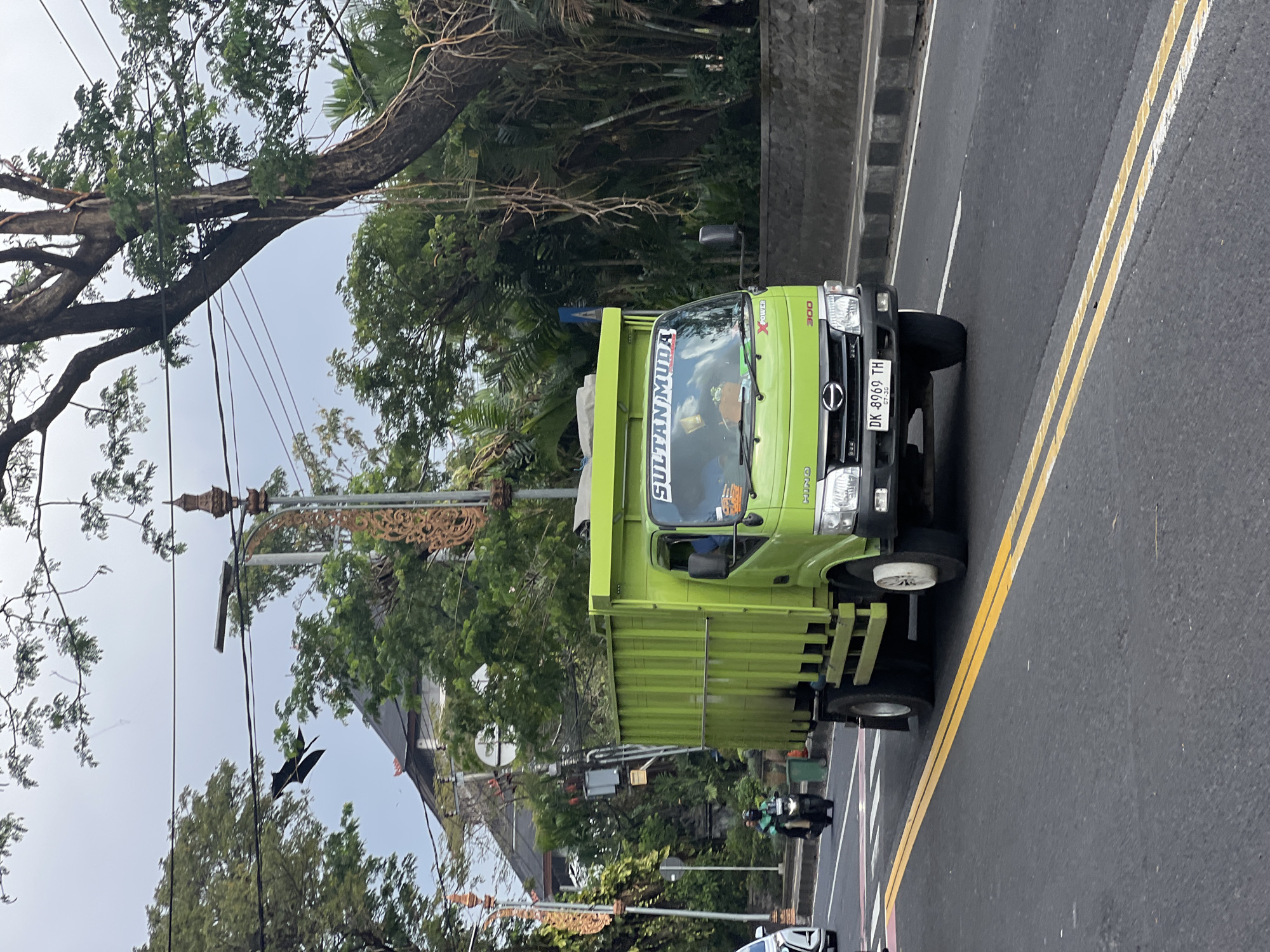 A green dump truck with "SULTAN MUDA" written on the windshield drives on a paved road in Denpasar, Indonesia. The truck has the license plate "DK 8969 TH." There is a person on a motorcycle with a passenger behind them, to the left of the truck. The background shows trees, a building with a red roof, and overhead power lines. The sky is overcast.
