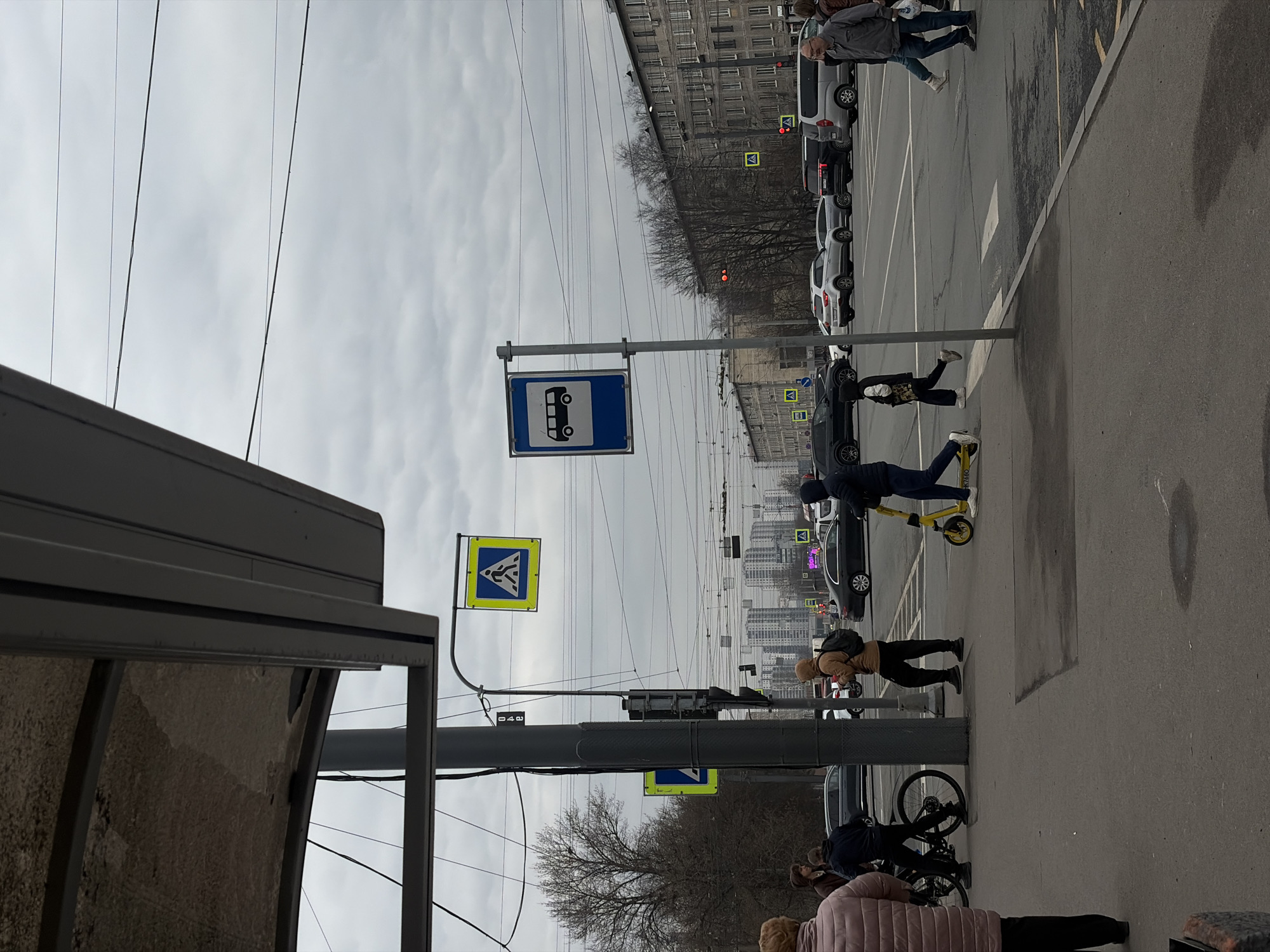 The image depicts a street scene in Saint Petersburg, Russia, under an overcast sky. A bus stop shelter is visible on the left. Two traffic signs, one with a bus symbol and the other with a pedestrian crossing symbol, are mounted on poles alongside the road. Several people are present: One person is riding a yellow scooter on the sidewalk, while others are walking or standing. A row of cars is parked along the street. Buildings are visible in the background.