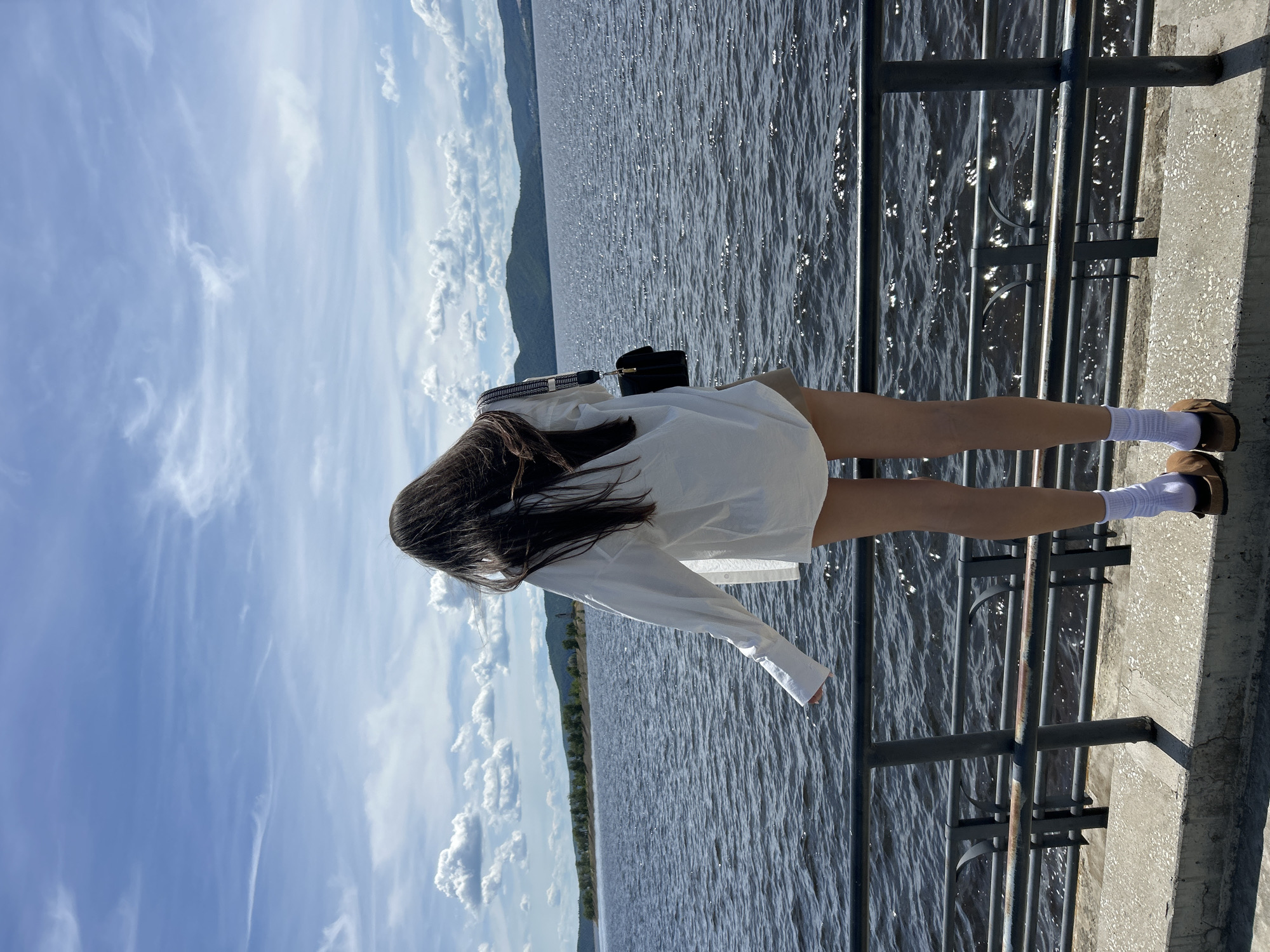 A rear view shows a person standing on a concrete ledge, facing a large body of water with a distant shoreline of hills under a blue sky with scattered clouds. The person has long dark hair, is wearing a loose white shirt, light-colored shorts, white socks, and brown sandals. A black purse with a patterned strap is slung over their right shoulder. A dark metal railing is in front of the person. The water is choppy and reflects sunlight. The scene is likely a waterfront promenade or park area in Tolyatti, Russia, possibly near the Zhigulevskoye Sea, a reservoir on the Volga River.