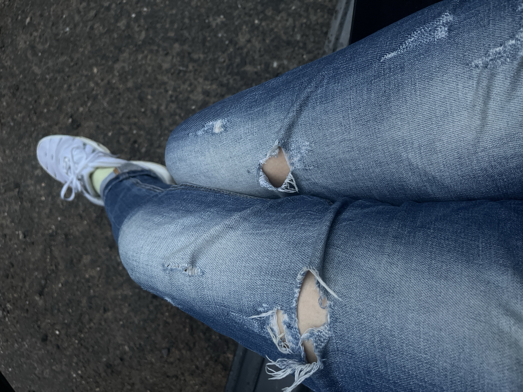 The image shows a pair of crossed legs wearing light-wash blue ripped jeans, with holes at the knees, and white sneakers. The left shoe has a visible light green sock. The legs are positioned over an area of dark grey pavement. The location in the city of Yablonovskiy, Russia is not possible to determine from the image.