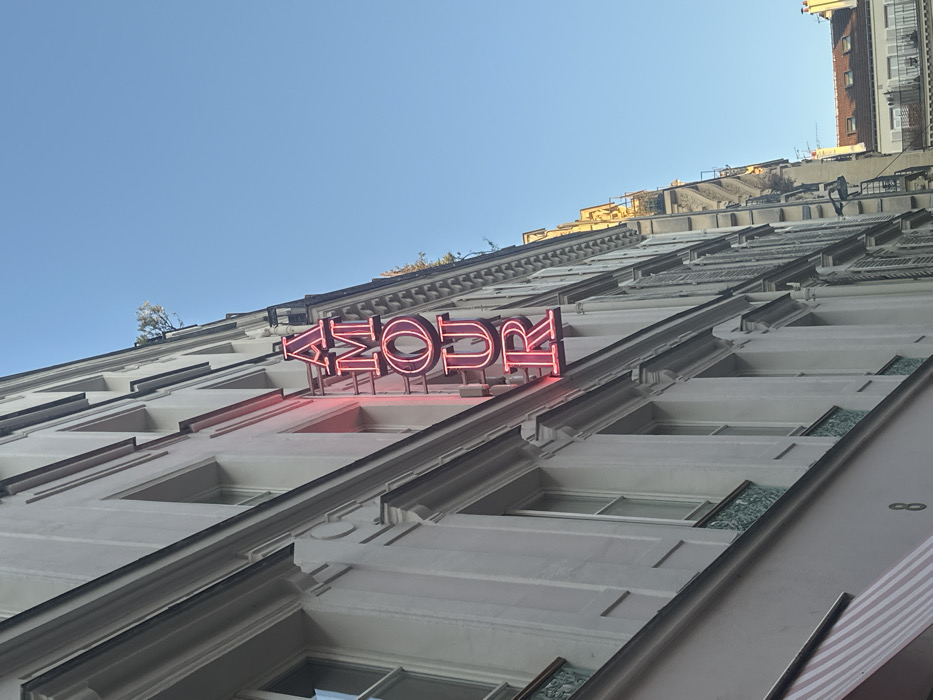 The image displays a clear, upward-angled view of a multi-story Parisian building facade under a bright blue sky. A prominent vertical neon sign, illuminated in red or pink, spells out "AMOUR" descending from top to bottom. The building features a light-colored, likely off-white, facade with several tall, narrow windows framed by decorative architectural elements. Below some lower-level windows, ornamental greenish-blue grilles are affixed. Partially visible at the bottom left is a pink and white striped awning, and the street number "8" is clearly discernible on the building's base. To the right, a continuous row of similar Haussmannian buildings extends into the distance, some with visible shutters. Distant rooftops and structures further down the street show areas of bright sunlight. The scene is devoid of people. This location is identified as the Hotel Amour at 8 Rue de Navarin, 9th arrondissement, Paris, France.