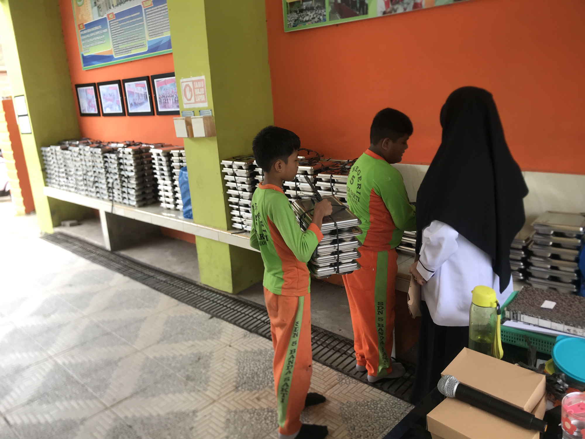 The image captures an indoor or semi-outdoor scene, likely within a school in Lhokseumawe, Indonesia. Three individuals are visible, two of whom are young boys or male students. Both boys are wearing matching uniforms consisting of lime green long-sleeved tops and orange track pants with a vertical green stripe. The text "SDN 5 BANDA SAKTI" is visible on the green stripe of the foreground boy's pants, identifying their school as State Elementary School 5 Banda Sakti, a district within Lhokseumawe. The other boy's shirt also shows "NEGERI 5".
The boys appear to be engaged in preparing or clearing up a meal service. The boy in the foreground is holding a stack of shiny, rectangular metal containers, possibly food warmers or serving trays, similar to the numerous stacks neatly arranged on a long, low shelf or counter to their left. The other boy, facing right, also seems to be interacting with these containers. A third person, partially visible on the far right, wears a black hijab and a white shirt, observing or assisting.
The setting features bright orange and lime green painted walls, creating a vibrant atmosphere. A large informational poster and four framed pictures depicting groups of people (likely students or staff) adorn the orange wall. A small sign on a green pillar clearly reads "TAMU WAJIB LAPOR," which translates to "GUESTS MUST REPORT," indicating a public or institutional building. On a table in the foreground right, a black microphone, two brown cardboard boxes, a green water bottle, and a patterned notebook are placed. The floor is tiled with a light-colored, geometric pattern, and a metal grate runs along the base of the counter. The overall impression is one of students actively participating in school-related tasks during what appears to be daytime.