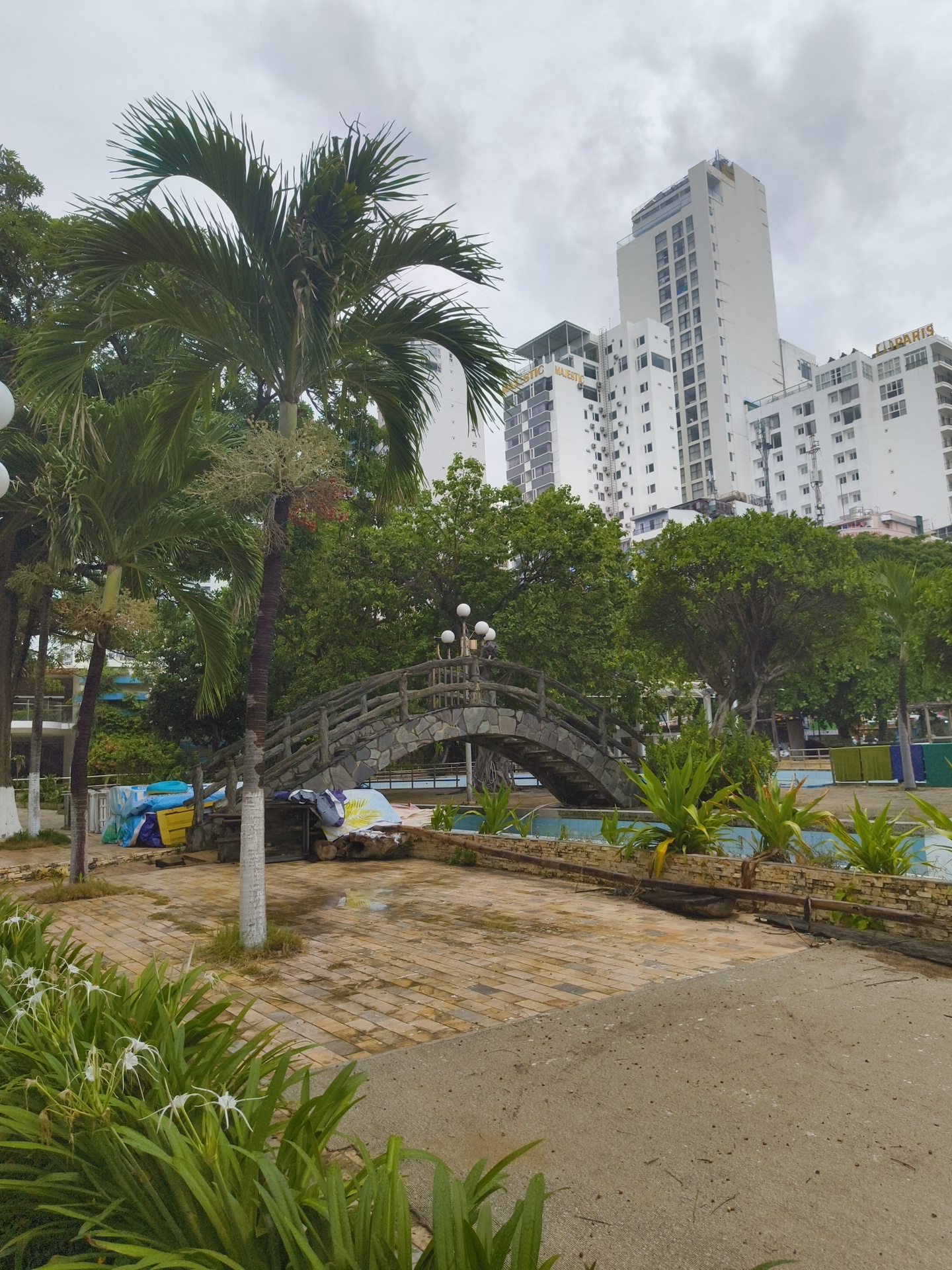 The image depicts a view of the Nha Trang beach area in Vietnam. In the foreground, a paved walkway with a yellow brick pattern is visible, leading towards a small stone bridge with wooden railings. White flowers and lush green foliage are present along the edge of the walkway. Behind the bridge, a swimming pool with blue water is partially visible, bordered by a stone wall and more greenery. Tall, modern white buildings, some adorned with gold lettering such as "MAJESTIC" and "DIAMOND," dominate the background, suggesting the presence of hotels or resorts. Several palm trees with their distinctive fronds are scattered throughout the scene. The sky is overcast with gray clouds. The scene appears to be an outdoor recreational area within a developed urban coastal environment. There are no people visible in the image.