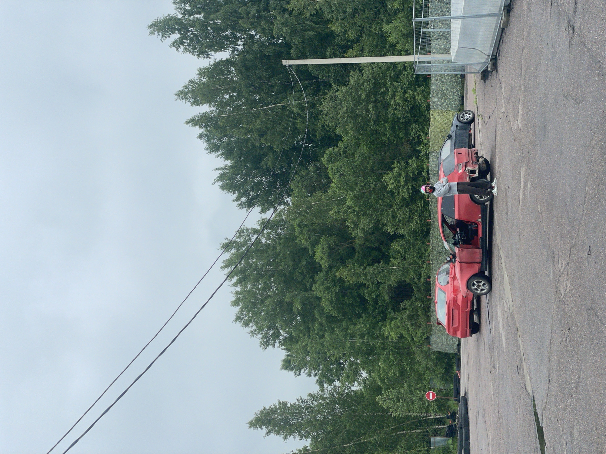 The image shows an outdoor scene in Ковалёво, Russia. The foreground is a cracked asphalt surface. In the middle ground, two red cars and a dark-colored car are parked. One red car is open, with its doors and trunk removed. A person wearing a pink helmet, a gray sweatshirt, and dark pants is standing next to the open red car, facing the camera. In the background, a dense line of green trees forms a backdrop under a gray, overcast sky. Power lines are visible stretching across the sky. To the right, a section of a metal fence with a white barrier is present. A red and white stop sign is visible to the left of the red cars.