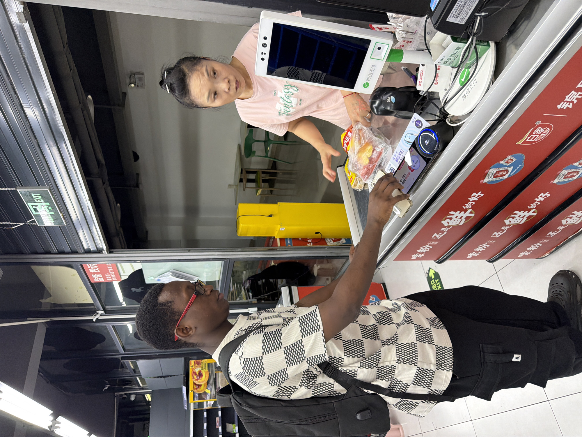 In the city of Jiangjin, China, a young Black man wearing a black backpack and a checkered t-shirt with red-rimmed glasses is at a supermarket checkout. He is holding a smartphone over a payment terminal, seemingly completing a transaction. A woman of East Asian descent, wearing a pink t-shirt, stands behind the counter, attending to the checkout. Various food items are visible on the conveyor belt. The environment appears to be a modern supermarket, with bright lighting and shelves visible in the background. A digital display screen and a scanner are part of the checkout counter. A yellow cabinet is also present behind the counter.