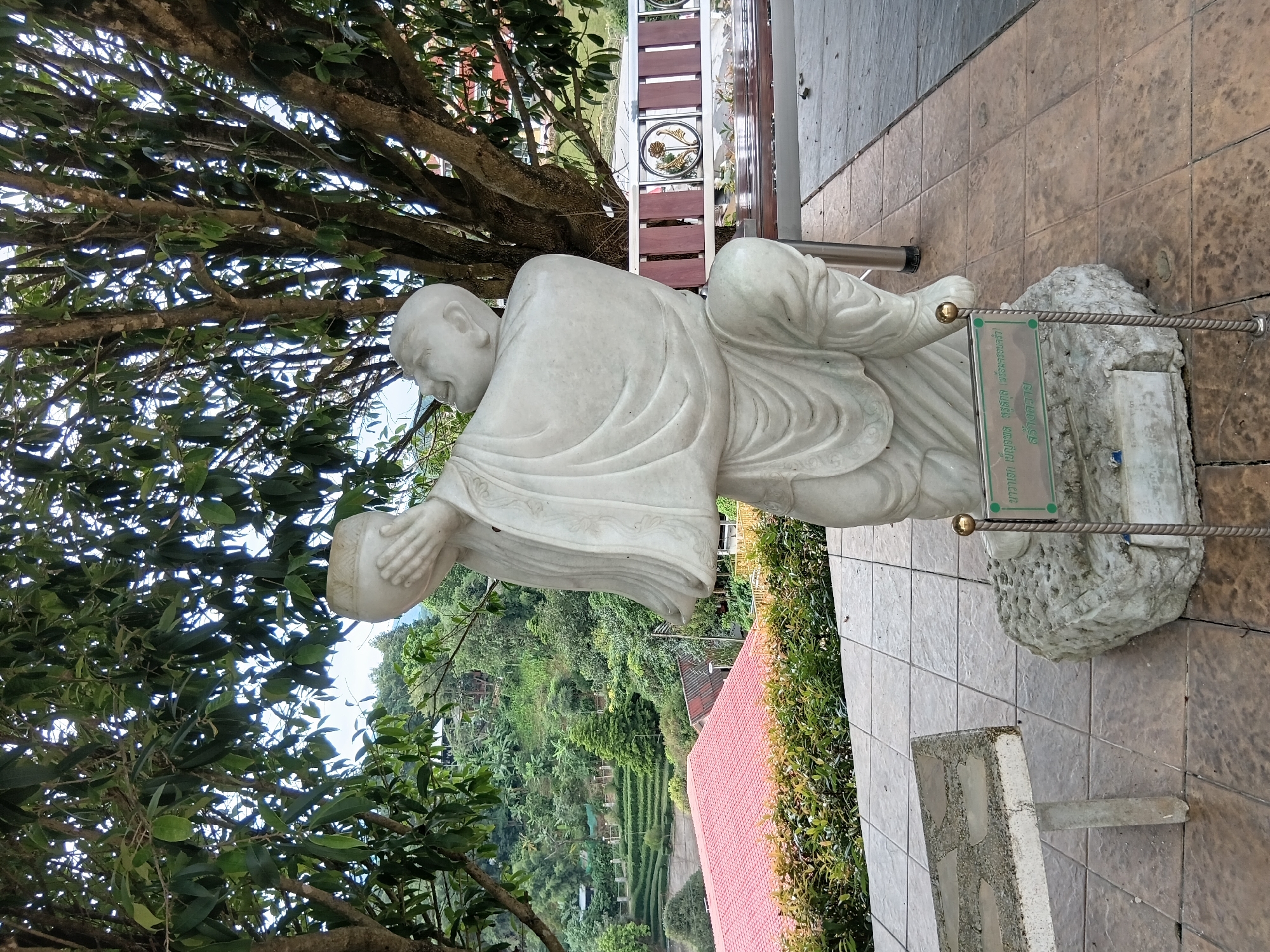A white marble statue of a bald, robed figure, possibly a monk, is positioned on a rough stone base. The figure is kneeling with its arms raised, holding a round, bowl-like object to its face. The statue is situated outdoors on a paved area, with a stone bench to the right and a low stone table to the left. In the background, a lush green hillside with terraced fields and buildings can be seen. A large tree with dense foliage partially shades the scene. A sign with green text is placed in front of the statue's base. The location is identified as บ้านเหล่าพัฒนา, Thailand.