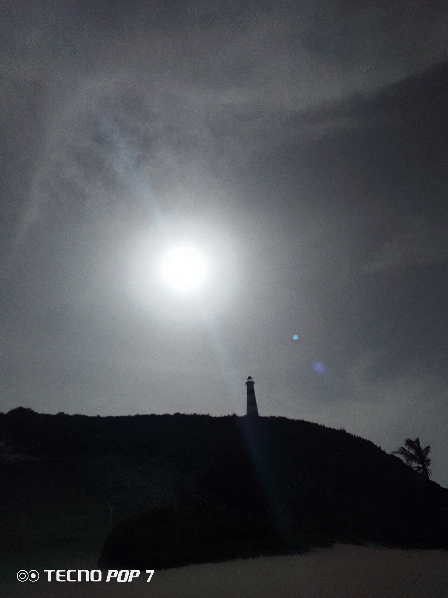 The image is a silhouette of a lighthouse atop a hill in Pedro González, Venezuela. The sky is overcast with the sun directly behind the lighthouse. There is vegetation on the hill and a palm tree on the right side of the hill. The foreground appears to be a beach. The image was taken with a TECNO POP 7 camera.