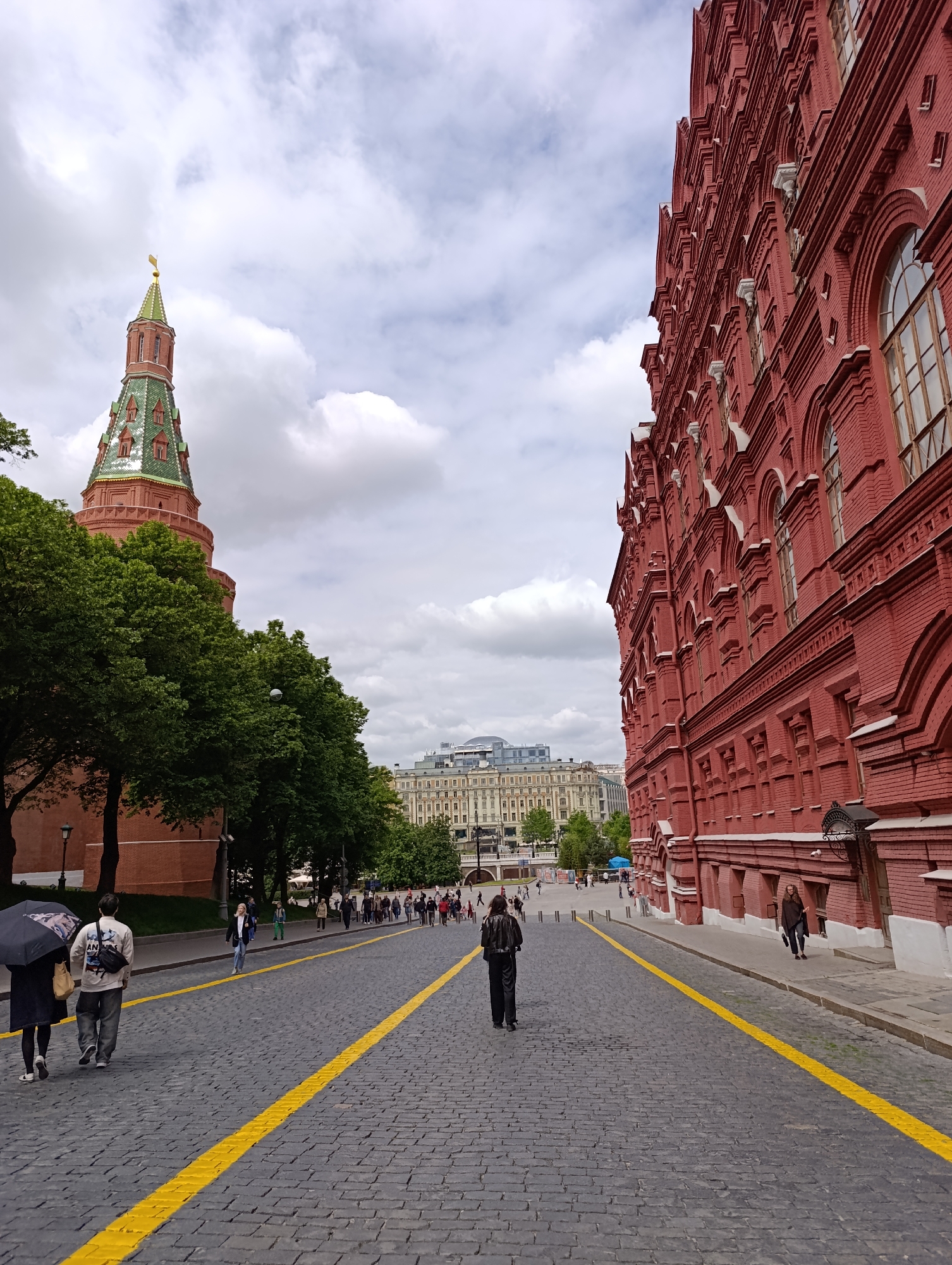 The image presents a wide-angle view of a historic urban street under an overcast sky. On the left, a tall, reddish-brown tower with a distinctive green, pointed roof and a golden spire rises above a line of lush green trees. This tower is identifiable as one of the Kremlin towers in Moscow, likely the Kutafya Tower.
On the right, a large, ornate red brick building with numerous arched windows and intricate architectural details dominates the frame. This structure is characteristic of the State Historical Museum, located adjacent to Red Square in Moscow. Its vibrant red facade stands out against the muted tones of the sky.
The street itself is paved with dark cobblestones and features two prominent parallel yellow lines running down its length, guiding pedestrians. Several people are visible walking along this path and on the sidewalks. In the foreground, two individuals walk away from the viewer; one carries a black umbrella. Further down, more pedestrians are scattered, some walking individually, others in small groups, suggesting a moderately busy area.
In the background, beyond the trees and the immediate street, lighter-colored buildings with classical designs are faintly visible under the cloudy sky. The overall scene depicts a famous pedestrian area in Moscow during cloudy daytime conditions. No discernible text is visible on any buildings or objects.