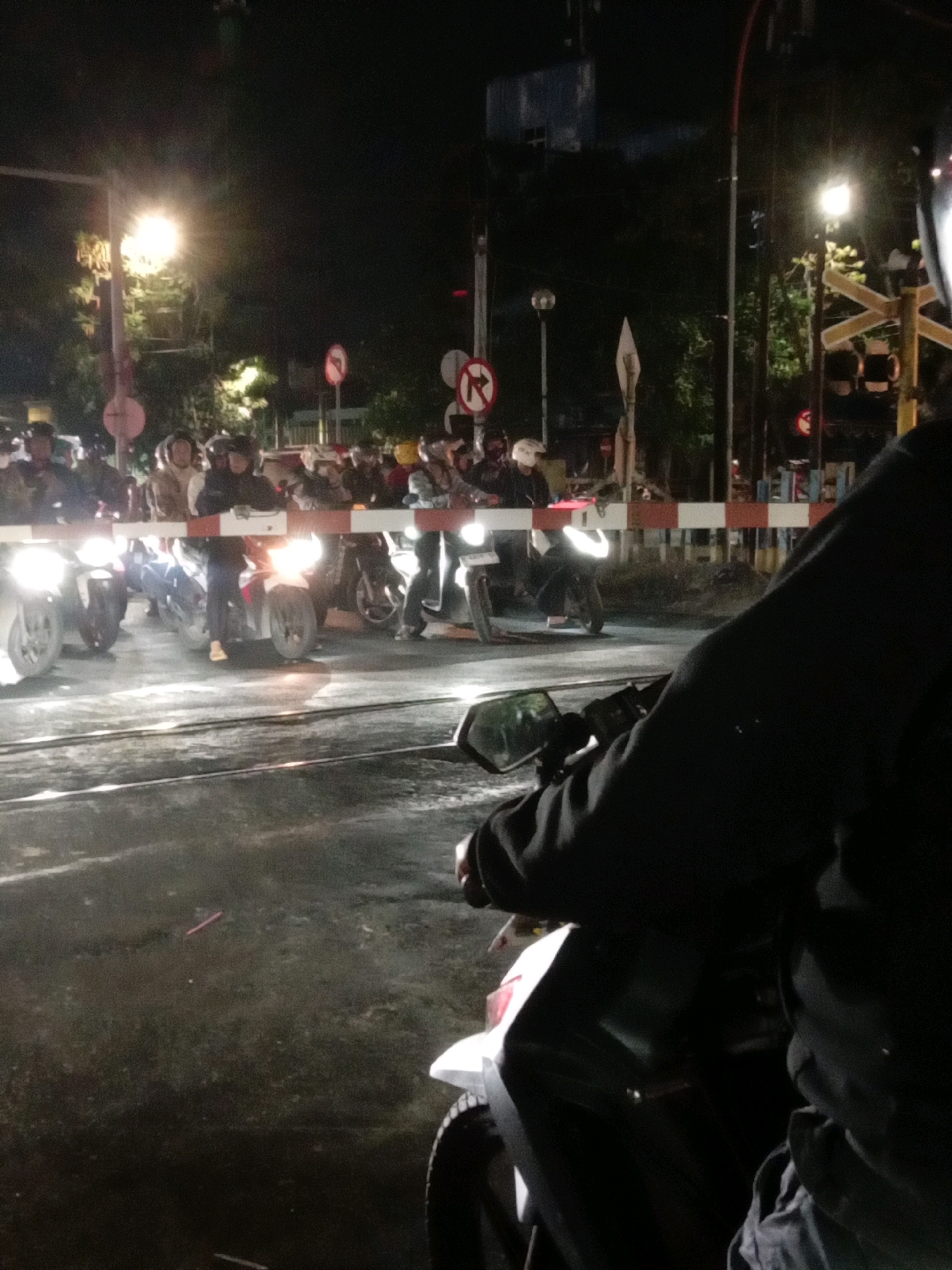 The image depicts a nighttime scene at a railway crossing in Surabaya, Indonesia. A group of motorcyclists are stopped at a red and white striped barrier, likely waiting for a train to pass. The area is illuminated by streetlights and the headlights of the motorcycles. In the foreground, the back of a person on a motorcycle is visible, looking towards the barrier. The environment appears to be urban, with some trees and buildings visible in the background. The overall atmosphere is one of a typical evening commute or travel interruption in a bustling Indonesian city.