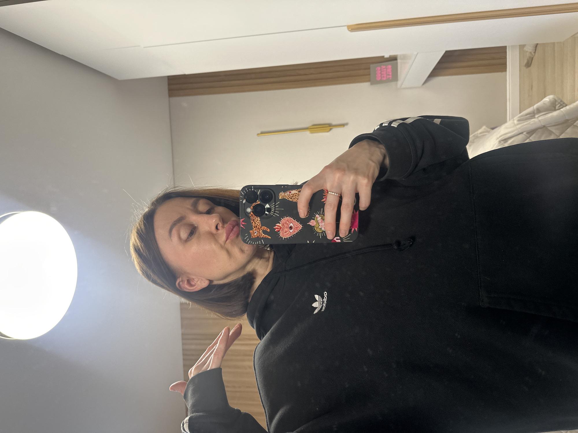 A woman is taking a selfie in a mirror. She is wearing a black Adidas hoodie and has a phone case with a colorful pattern. The background appears to be an interior space with a bed, light fixture, and a decorative arrow on the wall. The specific location in Votkinsk, Russia cannot be determined from this image.