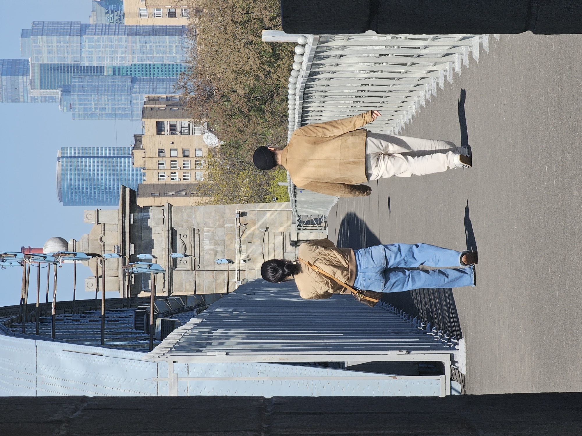 Here is a description of the image:
The image depicts two people walking on a bridge in an urban environment. One person is wearing a tan jacket and light-colored pants, along with a black beanie. The other person is wearing a tan jacket and blue jeans, with a brown purse hanging across their body. The bridge has a metal railing, and the cityscape of Moscow, Russia, including high-rise buildings, is visible in the background. The image is rotated.