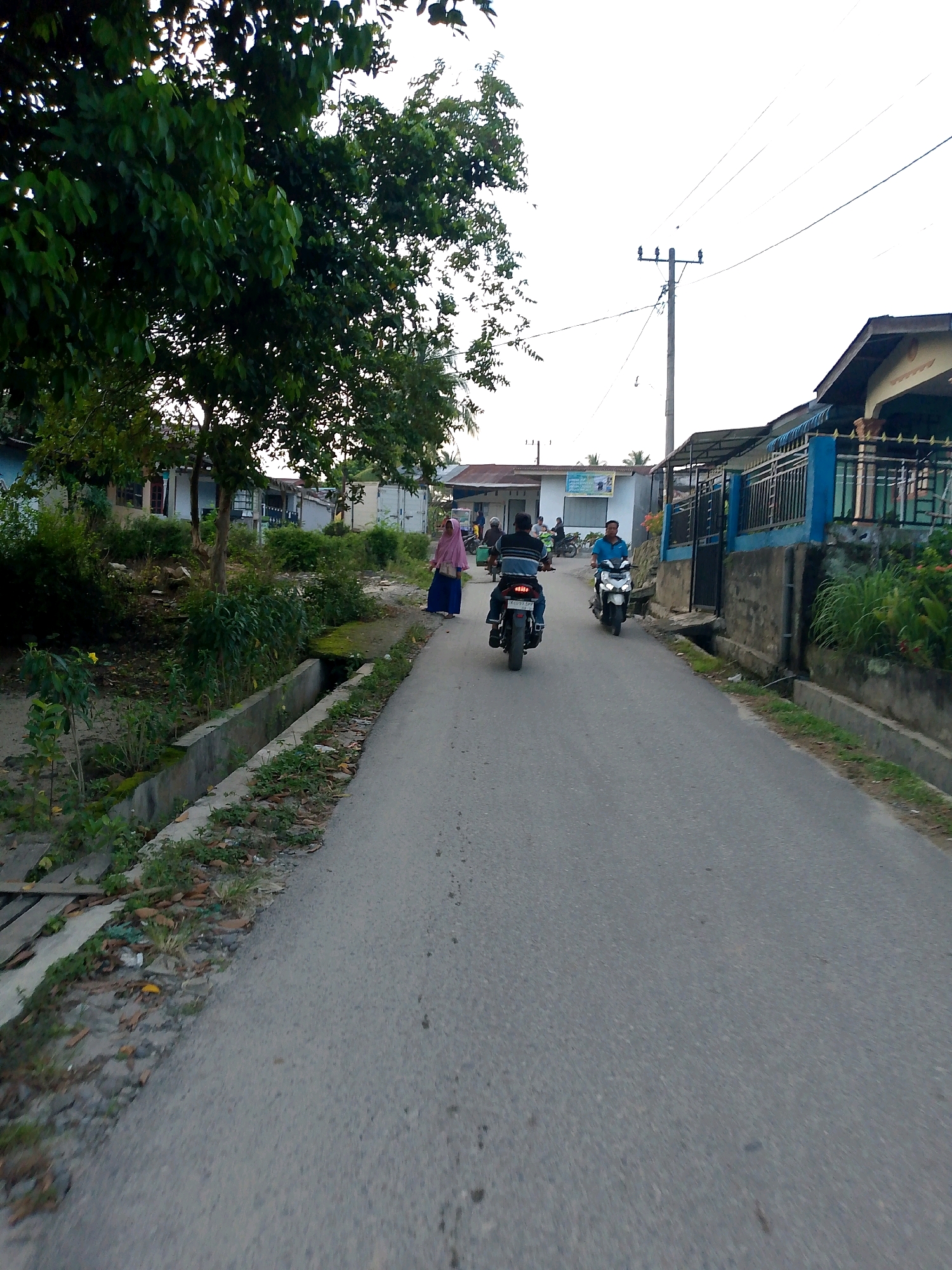 The image depicts a street scene in Dolok Merawan, Indonesia. A paved road runs through a residential area lined with houses and vegetation. Several individuals are present. In the foreground, the back of a person on a motorcycle is visible. Further down the road, two other individuals are riding motorcycles, one in blue attire and another in a blue shirt. On the left side of the road, a woman in a pink hijab and blue skirt is walking. The environment includes a concrete drainage channel along the side of the road, overgrown plants, trees, and utility poles with power lines overhead. The sky is overcast.