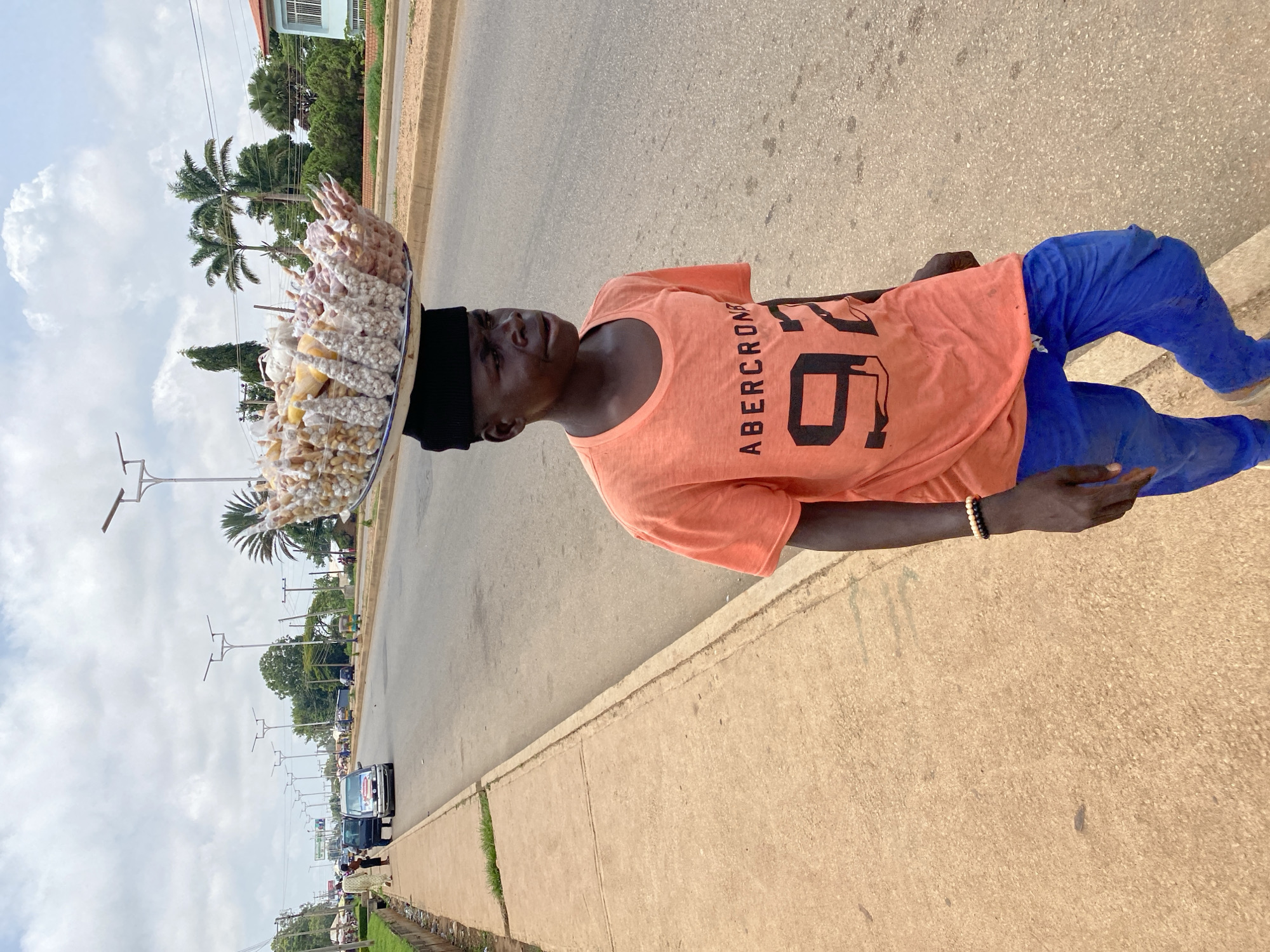 In Bukuru, Nigeria, a young boy walks along a paved sidewalk next to a road. He is wearing an orange t-shirt with the text "ABERCROMBIE 92" and blue pants. On his head, he balances a large, round tray filled with small, individually wrapped packages of food items, possibly snacks or candies, which are visible as light brown and white objects. The tray has a decorative rim. He is walking on the right side of the frame, with the road to his right and the sidewalk to his left. The road appears to be a city street with multiple lanes and is relatively busy with several cars and a tricycle visible in the background. Streetlights are positioned along the side of the road. Palm trees and other lush green vegetation are visible on the far side of the road, suggesting a tropical or sub-tropical environment. The sky is overcast with visible clouds. The overall scene depicts a common sight of street vendors in an urban setting in Nigeria.