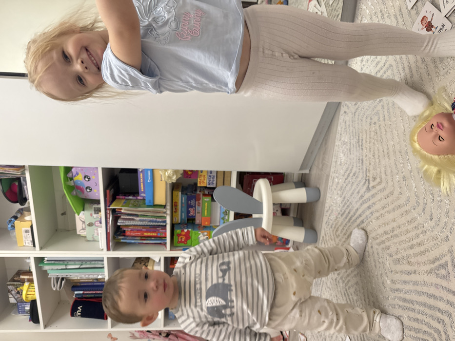 Two children are in the frame. One, a girl, is older with blonde hair, wearing a light blue top with the words "Funny Bunny" on it, and cream leggings. She is smiling at the camera. The other child, a younger boy, is wearing a striped shirt with an elephant design and cream pants. He is looking toward the left. Behind them is a white shelving unit filled with books and toys. On the floor is a doll with blonde hair and a grey and white patterned rug.