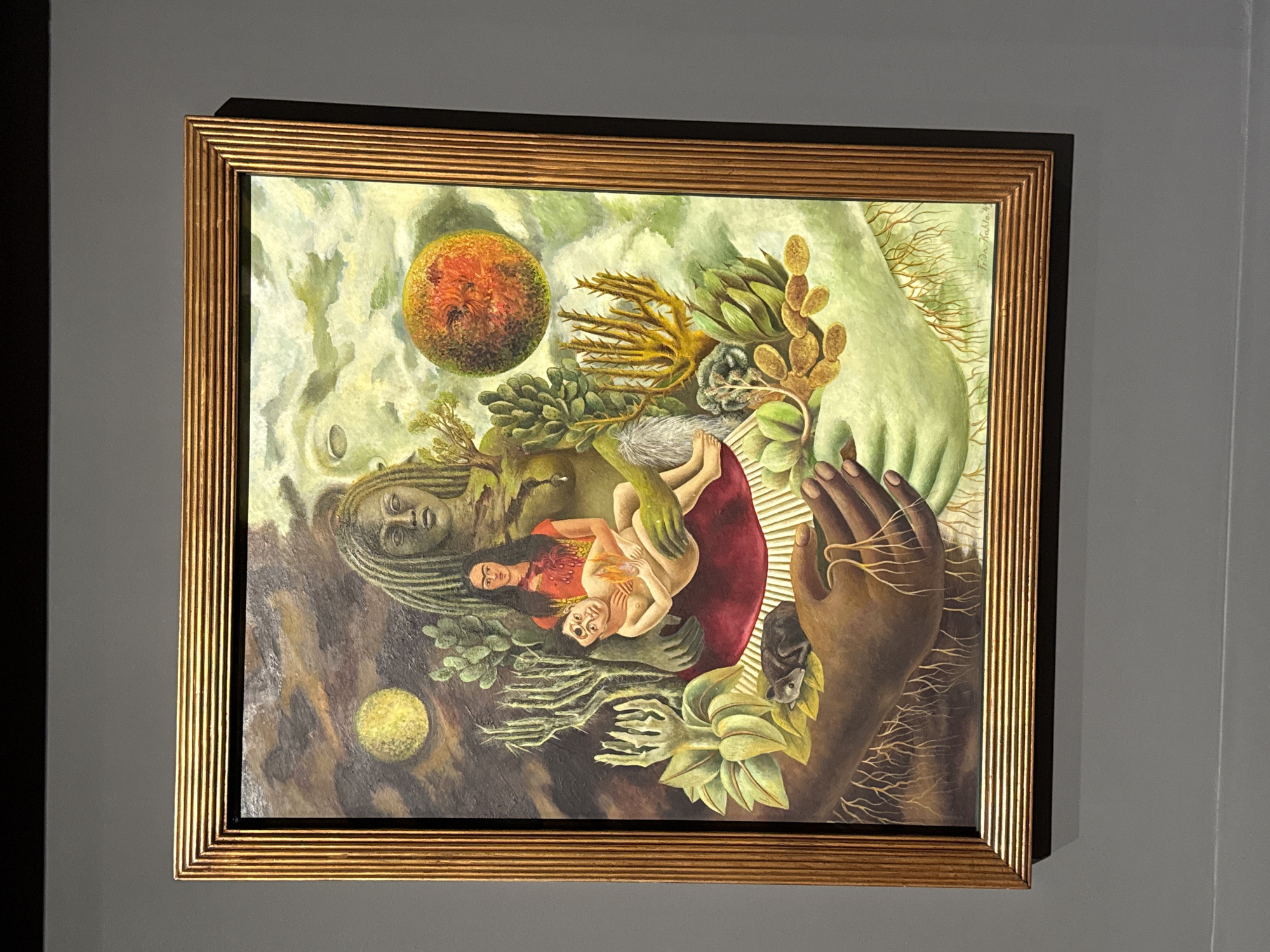 A framed painting hangs on a gray wall. The painting depicts a surreal scene with two figures cradled in a large hand. The sky is a mix of greens and whites. The Earth or a planet is represented by an orange orb in the top left. The painting seems to be Frida Kahlo's "Roots," and it is likely in the Museo Dolores Olmedo, Mexico City, Mexico.