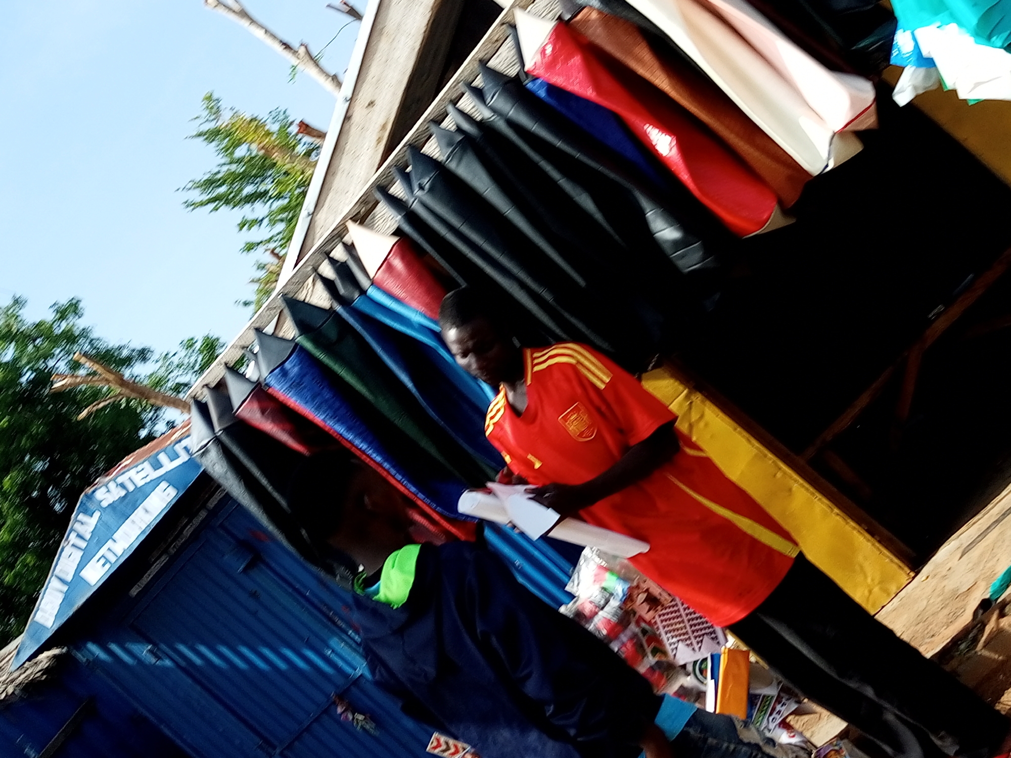 In Tempure, Nigeria, a vibrant street scene unfolds with a man in a red and yellow jersey holding a white object. He stands in front of a stall displaying a colorful array of what appear to be fabric rolls or folded materials, primarily in shades of black, blue, and red. To the left, a blue corrugated metal structure with signage reading "BUY DIGITAL SATELLITE NETWORKING" is visible. Another person, wearing a dark blue hooded jacket with a bright green collar, is partially visible in the foreground, facing away from the camera. The stall is constructed with wooden beams, and the backdrop suggests an outdoor setting with a clear blue sky and green foliage. The overall impression is one of a bustling marketplace, with goods neatly arranged for display and sale.