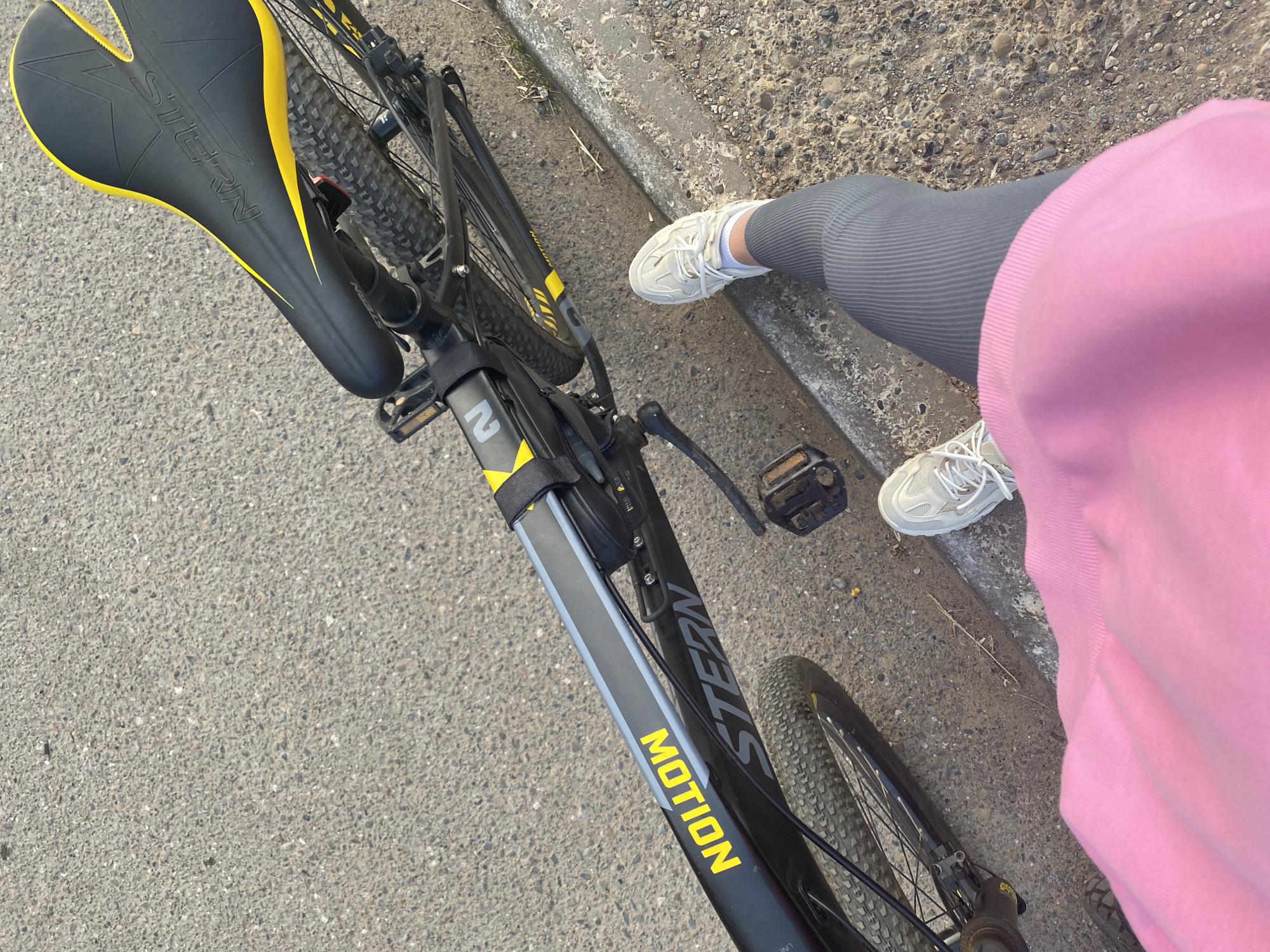 This media file captures a close-up perspective of a person standing next to a black bicycle with yellow accents. The bicycle features a "STERN MOTION" logo on its frame, and the seat is also black with yellow trim and the "STERN" brand name visible. The person is wearing grey ribbed leggings and white athletic shoes. A bright pink garment, possibly a hoodie or sweatshirt, is also visible, draped over the person's legs and obscuring part of the view. The bicycle is positioned on an asphalt surface, likely a road or path, adjacent to a concrete curb. The setting appears to be outdoors, with a slightly weathered and granular texture to the ground and curb. The lighting suggests it might be daytime, possibly late afternoon due to the angle of the shadows, though the sky is not visible. There are no clear indicators of specific weather conditions, but the scene has a casual, everyday feel. The image is taken from a high-angle perspective, looking down at the bicycle and the person's legs and feet. The overall mood is neutral, focusing on the elements of the scene and the objects within it.