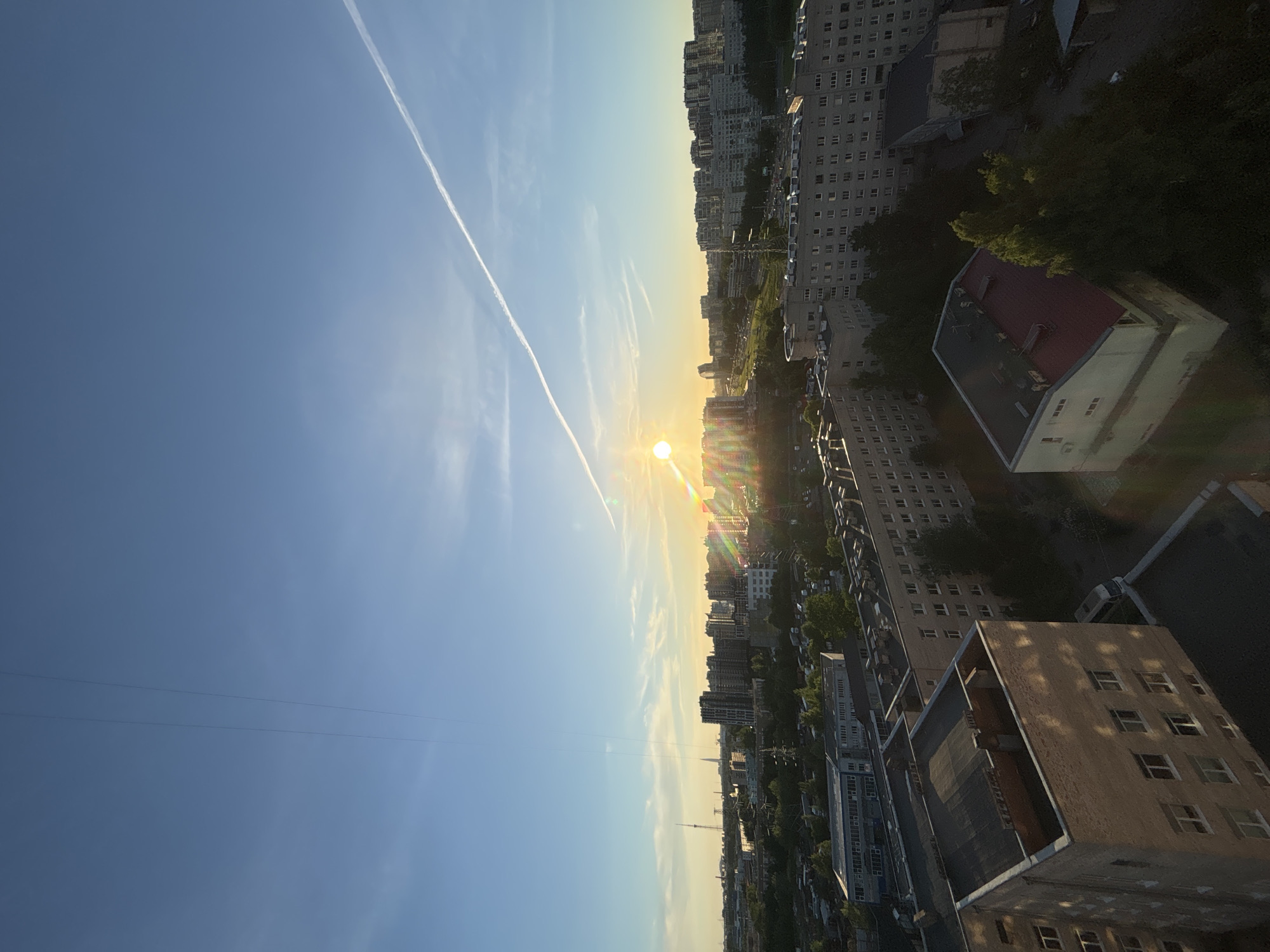 This is an aerial shot looking out over a city at sunset. The sky is a clear blue with a few wispy clouds. The sun is setting on the horizon, casting a warm glow over the scene. In the foreground, there are several multi-story apartment buildings with flat roofs. The buildings are made of brick and concrete, and many of them have a similar, utilitarian design. Trees are interspersed among the buildings, and there are some roads and vehicles visible. In the distance, more apartment buildings and a few taller structures, including what appears to be a radio tower, can be seen. A contrail from an airplane stretches across the sky. The lighting suggests it is late afternoon or early evening.