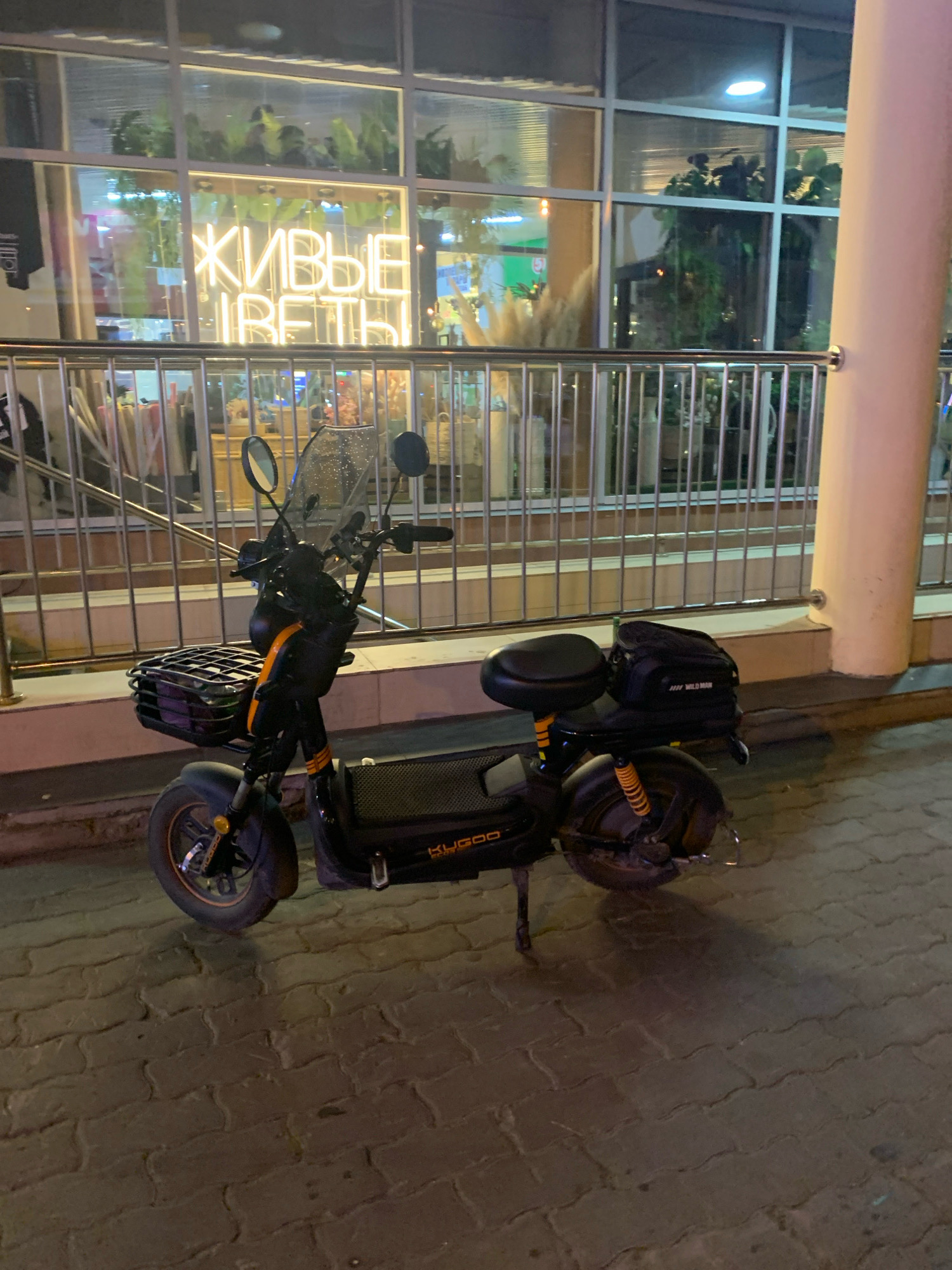 An outdoor, medium shot captures a black electric scooter parked on a paved walkway at night. The scooter, a Kugoo model, is positioned slightly angled towards the left, with its front wheel on the left and the rear wheel on the right. It features a black seat, a basket on the front, and a small black bag mounted behind the seat. Yellow accents are visible on the scooter, including on the suspension springs and stripes on the front fork.
In the background, a building with large glass windows is illuminated, displaying an interior with plants and neon signs. One sign, in Cyrillic script and glowing yellow, reads "ЖИВЫЕ ЦВЕТЫ" which translates to "LIVE FLOWERS". A metal railing runs horizontally across the middle ground, separating the walkway from the building's facade. The walkway itself is constructed of interlocking pavers, creating a patterned surface. The scene appears to be in a commercial or urban area, given the building and street lighting. The overall lighting suggests it is nighttime. The specific location of Arkhangelsk, Russia, is provided as context.
