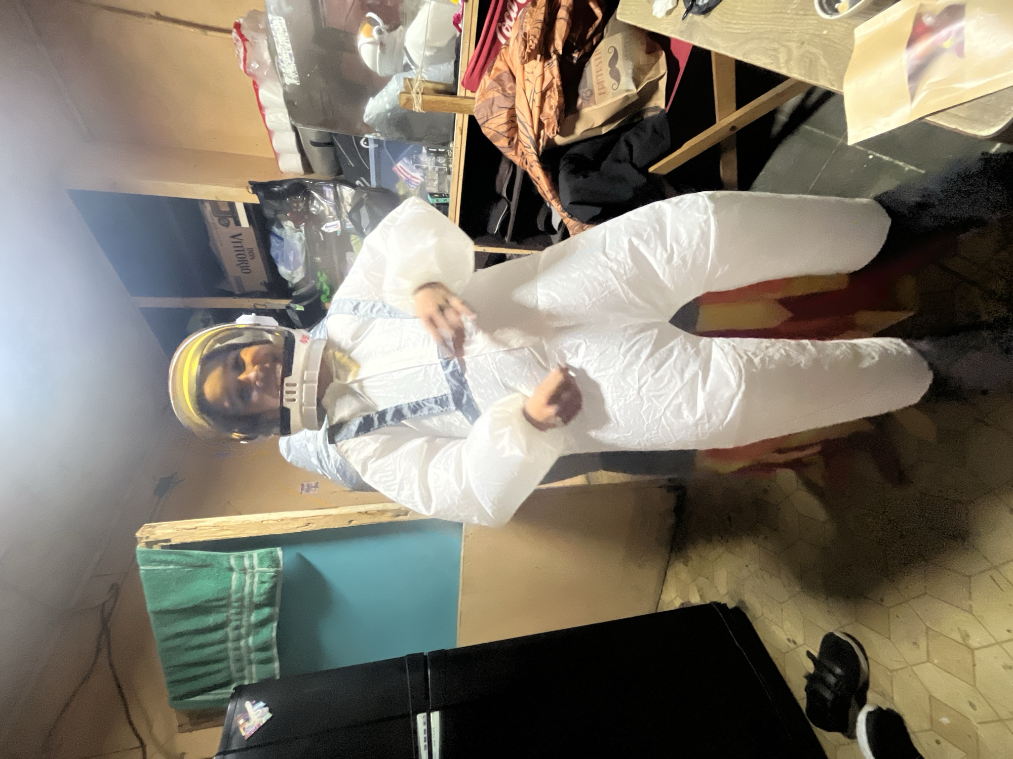 The image depicts a person inside a room wearing a white astronaut costume. The person is standing and appears to be posing or moving their arms. The costume is bulky and covers the entire body, with a helmet that has a yellow visor and a white faceplate. The room appears cluttered, with various items visible in the background, including shelves filled with objects, clothing, and what looks like a refrigerator on the left side of the frame. The walls are painted in neutral colors, and the floor is tiled. There are no clear indicators of the city of Iquique, Chile within the image.