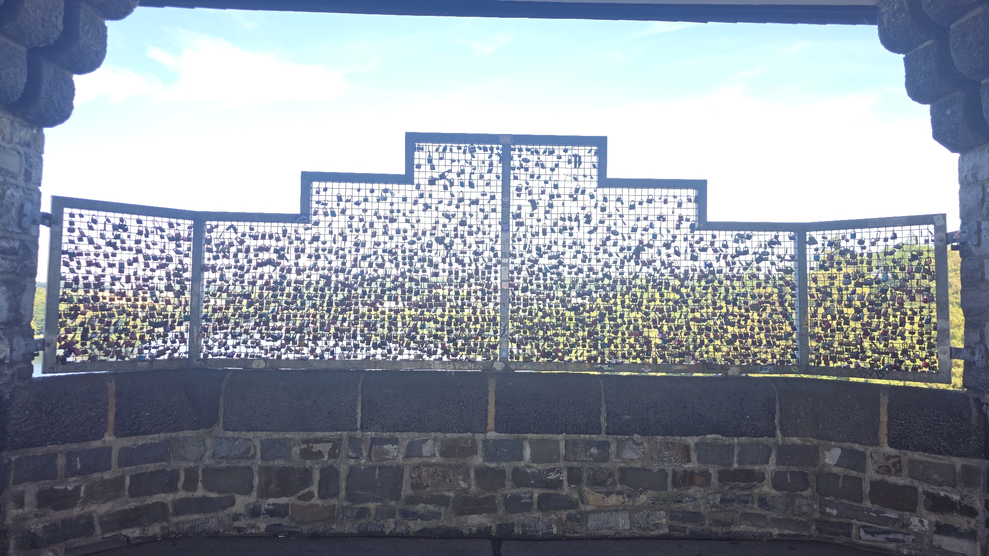 The image shows a viewpoint overlooking a body of water, likely the Möhnesee in Germany. The foreground features a stone wall and a metal railing covered in numerous padlocks. The padlocks are of various colors and sizes, densely covering the mesh of the railing, suggesting a tradition of attaching them as symbols of love or commitment. Beyond the railing, the water is visible, and on the far bank, a treed landscape is present. The sky above is bright with some clouds. The overall scene suggests a popular tourist or romantic spot.