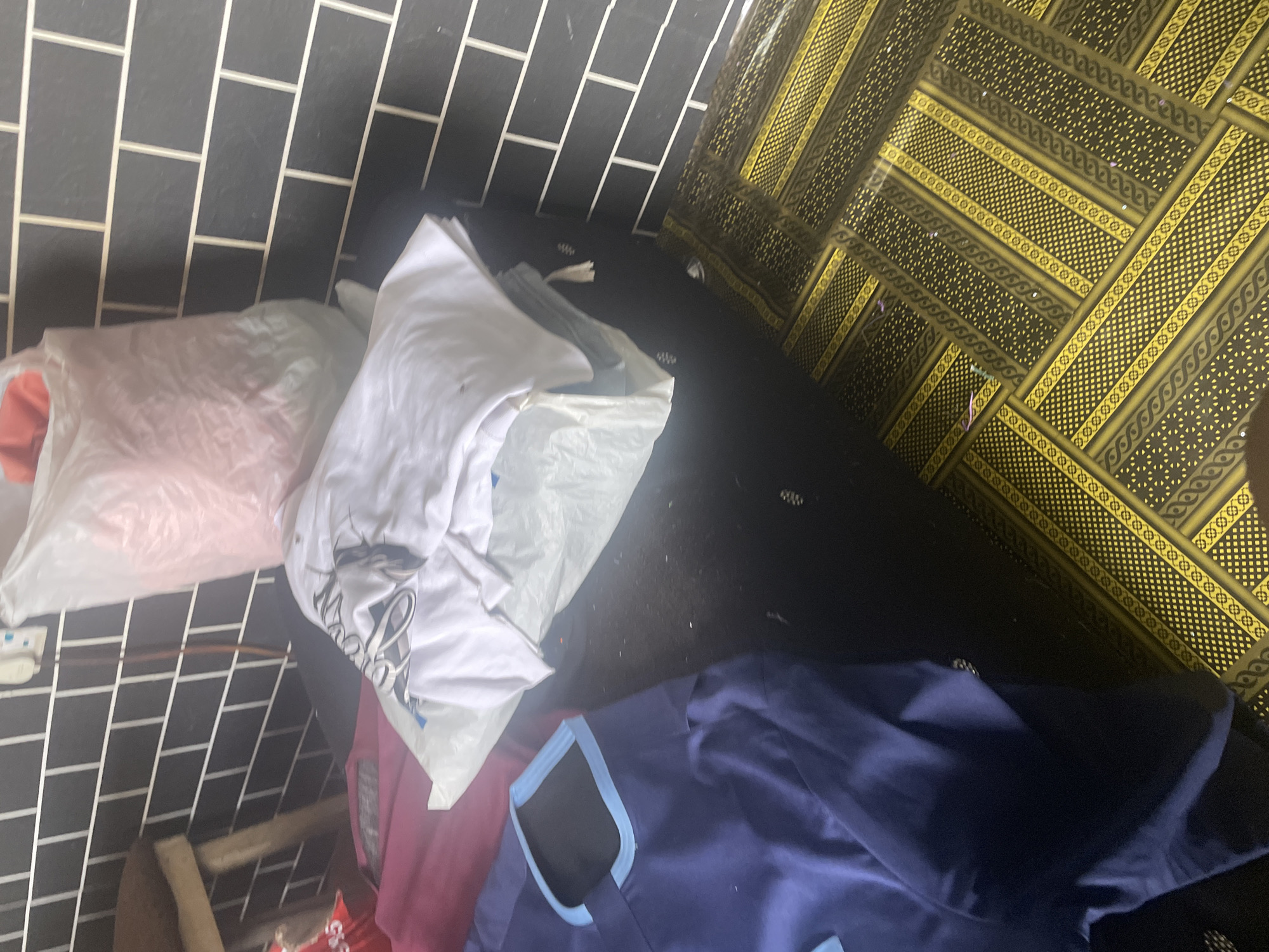 In Jauro Yinu, Nigeria, a scene unfolds within what appears to be a domestic setting, possibly a laundry area or storage space. The background features a dark grey tiled wall with white grout lines, arranged in a brick pattern. To the left, a wooden stool or bench is partially visible, with a dark cushion on top.
Dominating the foreground are piles of clothing. A large, translucent plastic bag filled with pink material is placed on a surface. Next to it, a white t-shirt with black lettering is folded and lying on top of more plastic-wrapped items. A navy blue garment, possibly a uniform or scrub top, with a distinctive light blue trim around the neckline and edges, is spread out in the lower left quadrant of the image. The floor is covered with a black rug or carpet, overlaid with a patterned yellow and black mat. The lighting suggests an indoor environment, with shadows cast by the objects. The overall impression is one of organized clutter, with laundry or textiles being processed or stored.