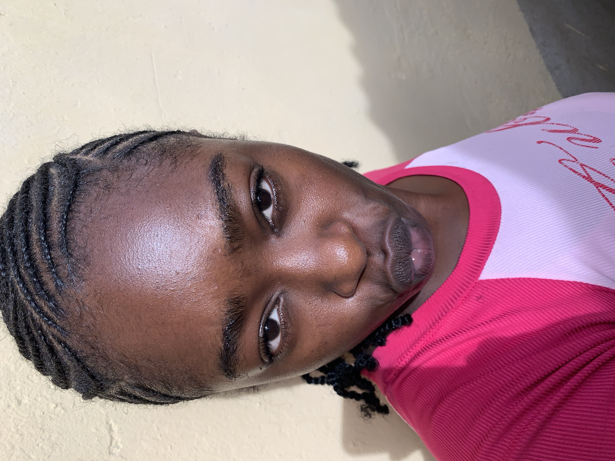 A close-up, slightly angled shot depicts a woman of African descent with cornrow braids. She is wearing a pink and white ribbed top. The background is a plain, light yellow wall. The location is Rafin Par, Nigeria.