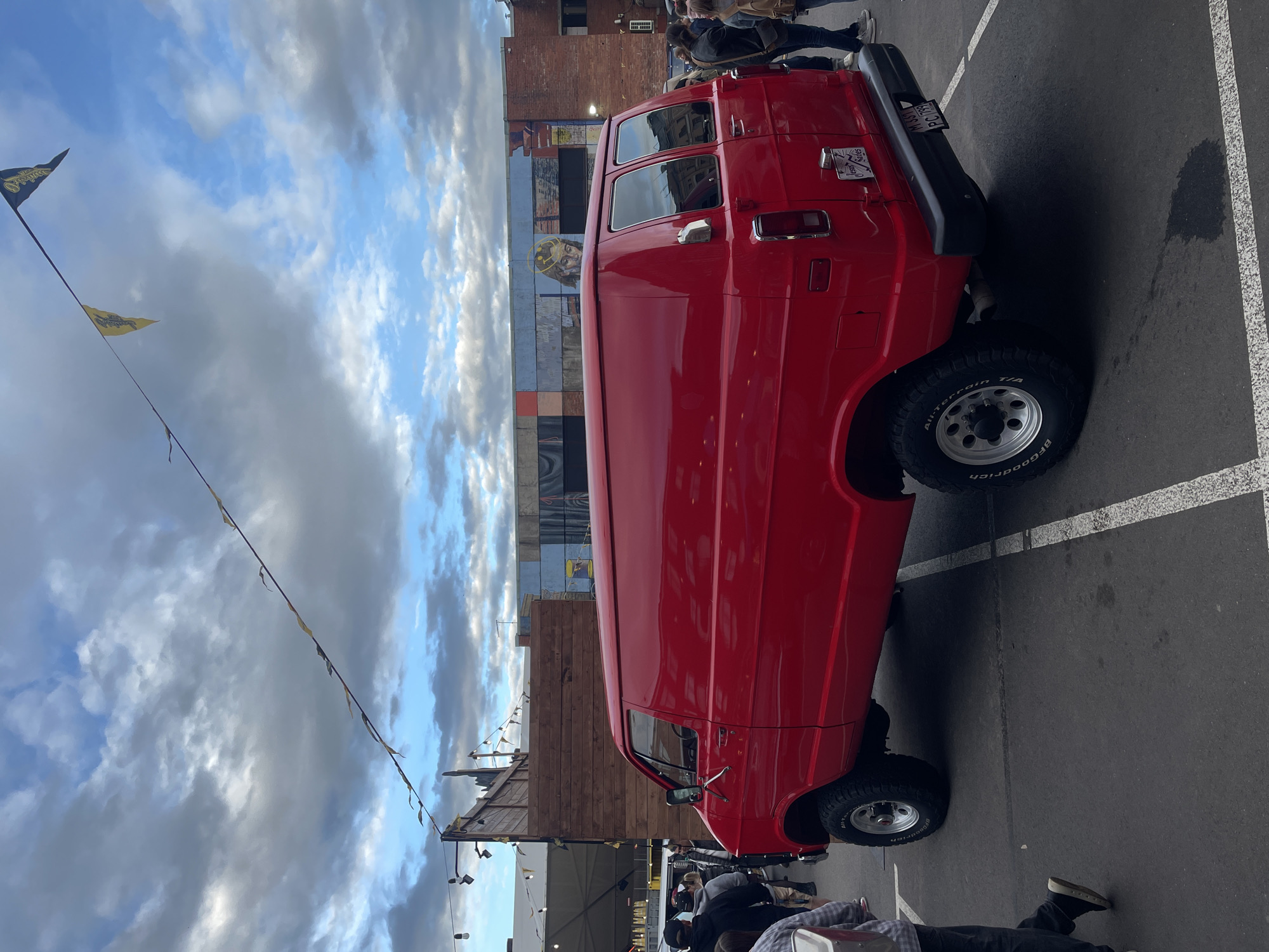 A red, lifted van is parked on asphalt with white parking lines. The van has large, off-road tires and a custom license plate on the back. In the background, a mural of a person with a halo adorns a building. String lights and small flags are visible overhead. Several people are milling about in the background, some walking and others standing. The sky is cloudy. The scene is not in Saint Petersburg, Russia.