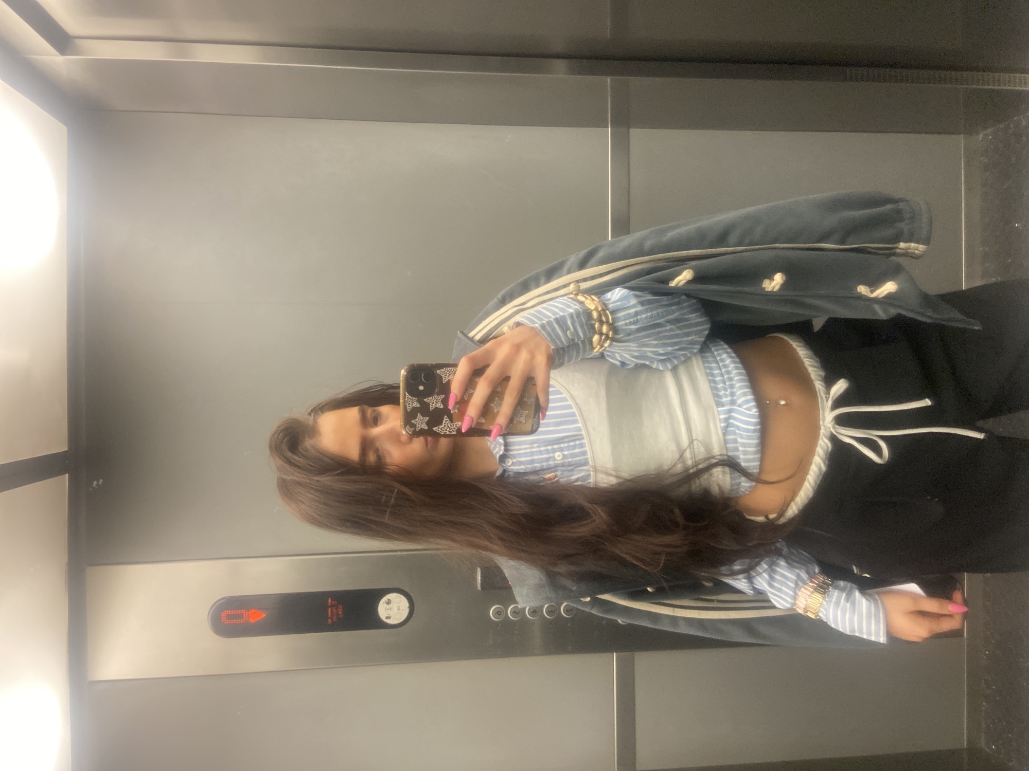 A young woman stands in an elevator, taking a selfie with her smartphone. She has long, brown hair and is wearing a blue and white striped shirt, a white corset, and black pants. She is also wearing gold bracelets and has pink nail polish. A denim jacket hangs off her shoulder. The elevator has a metal interior with elevator buttons visible. The location and city are not discernible from the provided image.