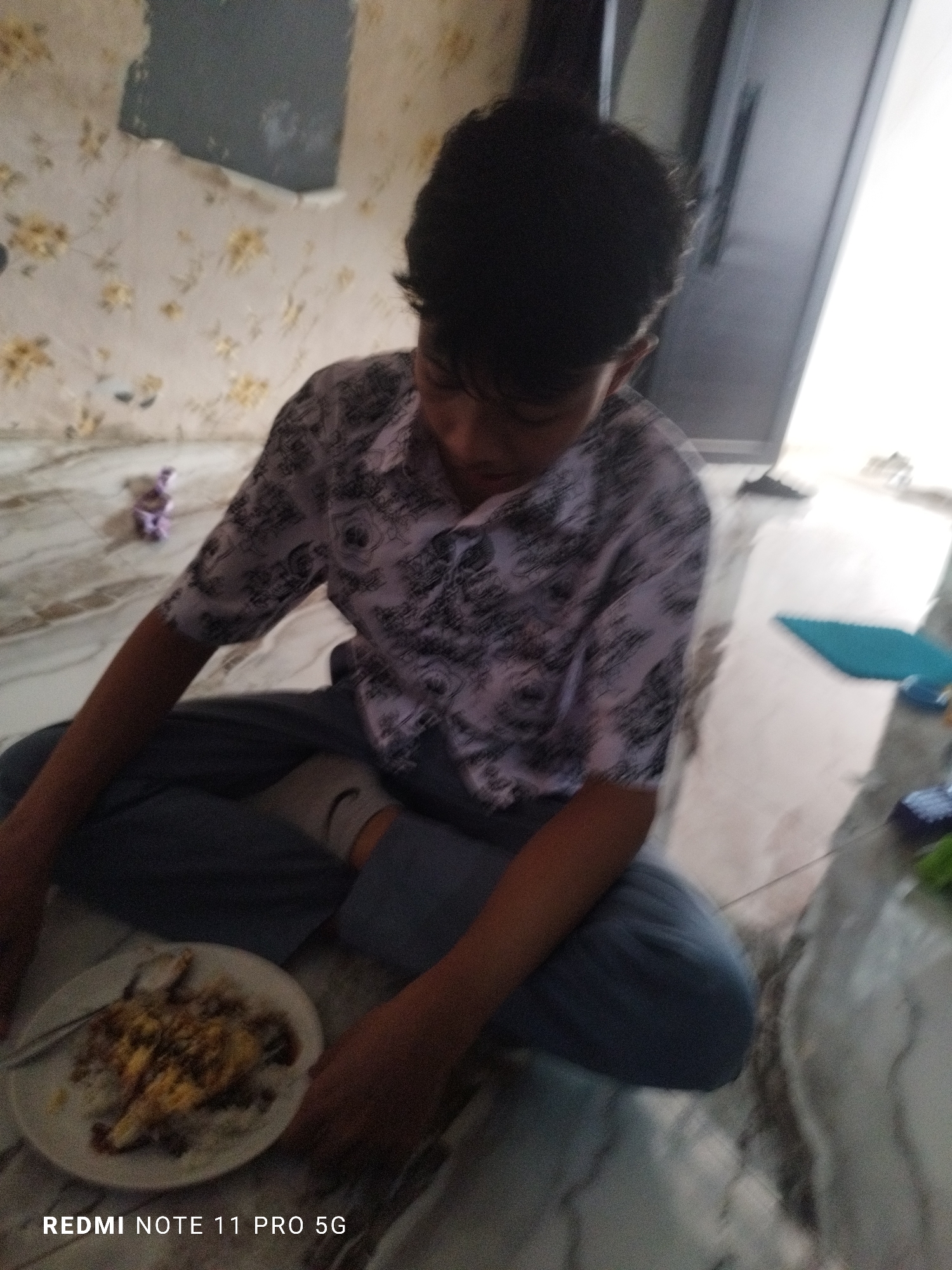 A young male, appearing to be of East Asian descent, is seated on a marble floor. He is wearing a short-sleeved collared shirt with a dark floral pattern and gray pants. His hair is dark and styled in a way that covers part of his forehead. In front of him is a white plate containing a meal, which appears to be rice topped with some kind of meat or protein. A spoon rests on the plate. The background consists of a wall with a floral wallpaper pattern, some of which is peeling or damaged, and a doorway or closet door. The lighting suggests an indoor setting, possibly a home. The image also displays a "REDMI NOTE 11 PRO 5G" watermark in the bottom left corner. The overall scene is somewhat blurred, giving a candid and informal impression. The location is identified as Kaduhejo, Indonesia.