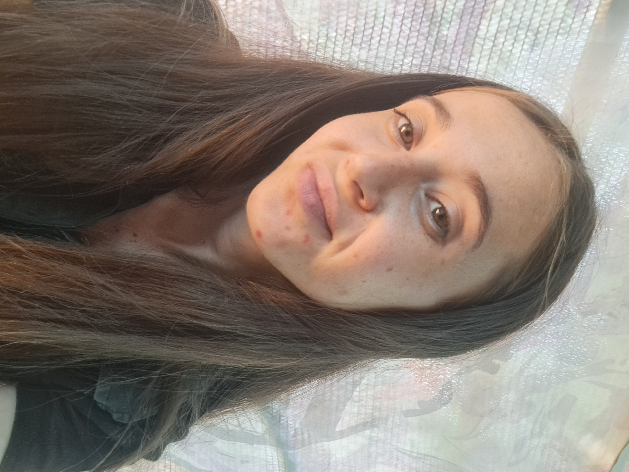 A young woman with long, dark brown hair poses for a selfie. She is looking directly at the camera with a slight smile. Her face is lightly freckled, and she has a few blemishes on her chin. She is wearing a dark-colored shirt. The background appears to be a window with a sheer curtain. The image was taken in Vypasne, Ukraine.