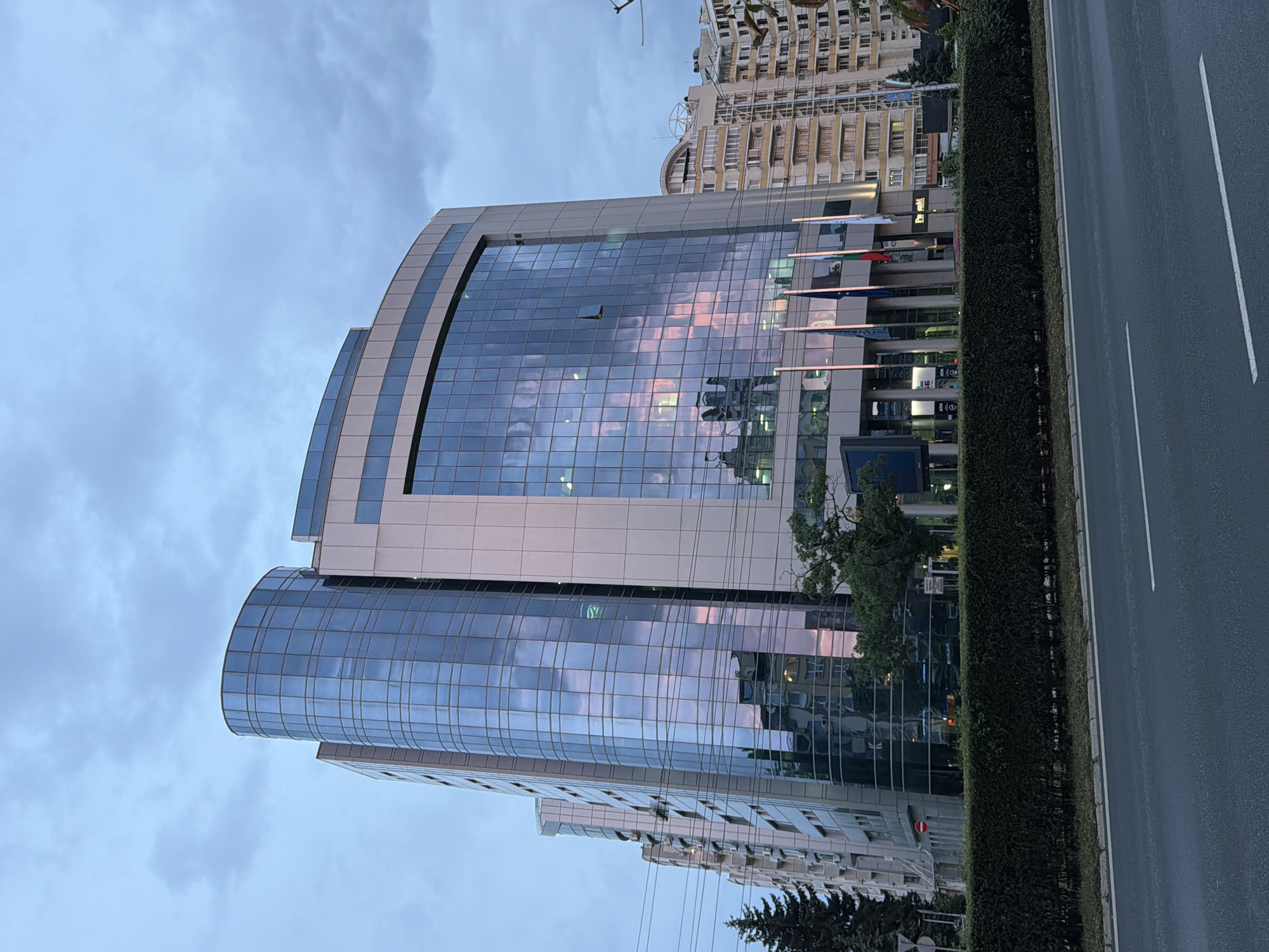 The image shows a modern office building with a curved glass facade in Sofia, Bulgaria. The building is several stories tall and reflects the sky, which is cloudy with pink and orange hues, suggesting it is either sunrise or sunset. The facade is a combination of glass panels and lighter-colored material. In front of the building, there are several flags, and a row of manicured bushes. To the left of the main building, another building with a similar architectural style is partially visible. Further to the right, a taller, older residential building with multiple balconies is seen. A tree is positioned in front of the main building, and some street lights are visible. A road with lane markings runs in the foreground, indicating the building is located in an urban area. The overall scene depicts a city view during twilight hours.