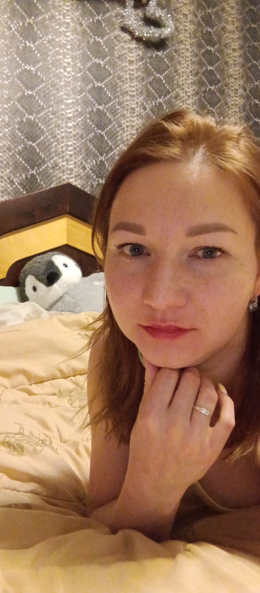 A woman with reddish-blonde hair is lying on a bed with a beige duvet and headboard. Her head rests on her hand, and she is wearing a silver ring. Behind her is a stuffed penguin toy. The background is a patterned wall covering. The location is unknown.