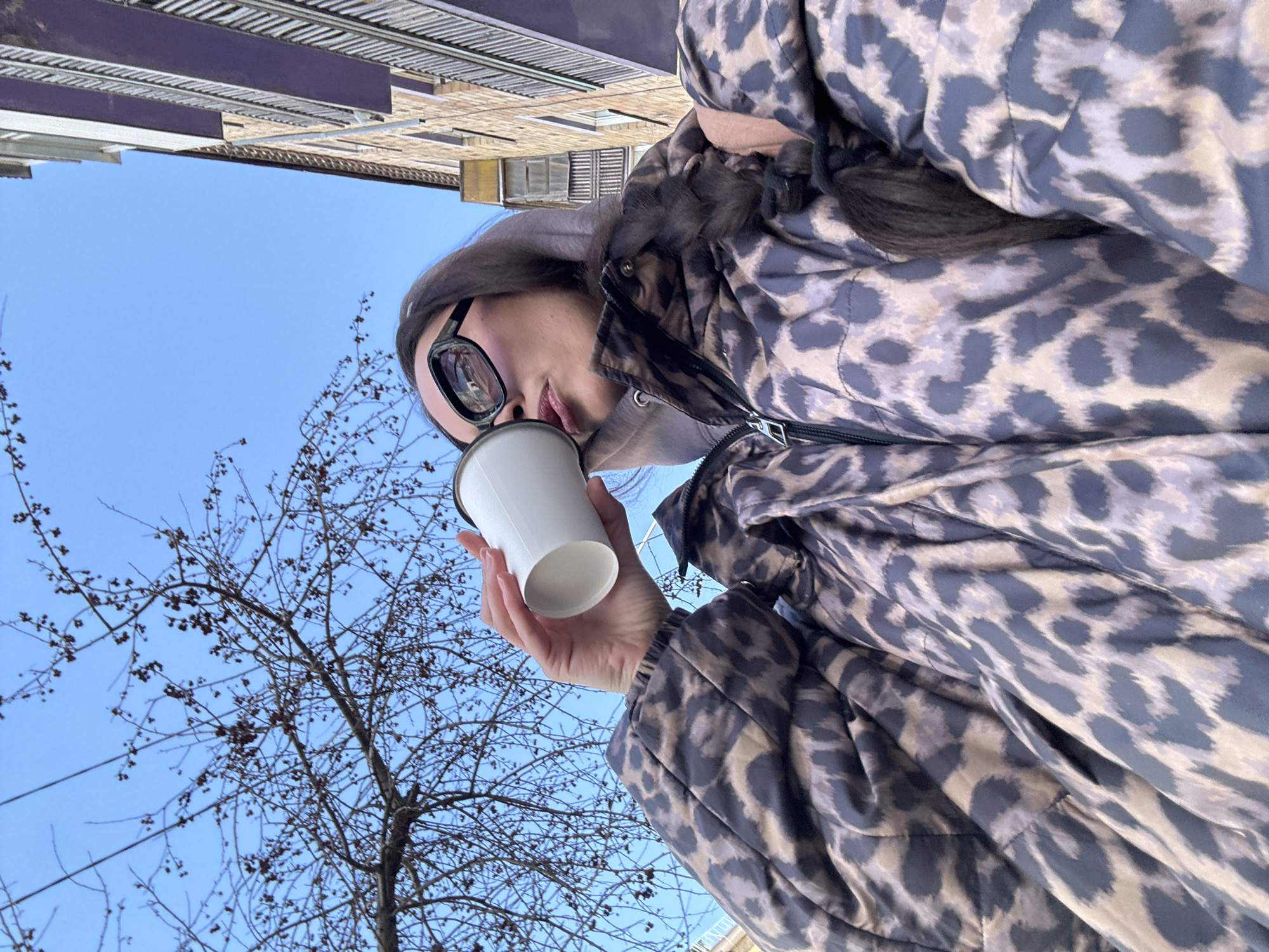 A woman is drinking from a white cup, likely coffee, outdoors. She's wearing a patterned puffer jacket and glasses, and the photo is taken from a low angle looking up. A bare tree is visible against a blue sky. Above, a multi-story brick building with metal and white accents is visible. It's difficult to pinpoint the exact location in Krasnoyarsk without additional landmarks.