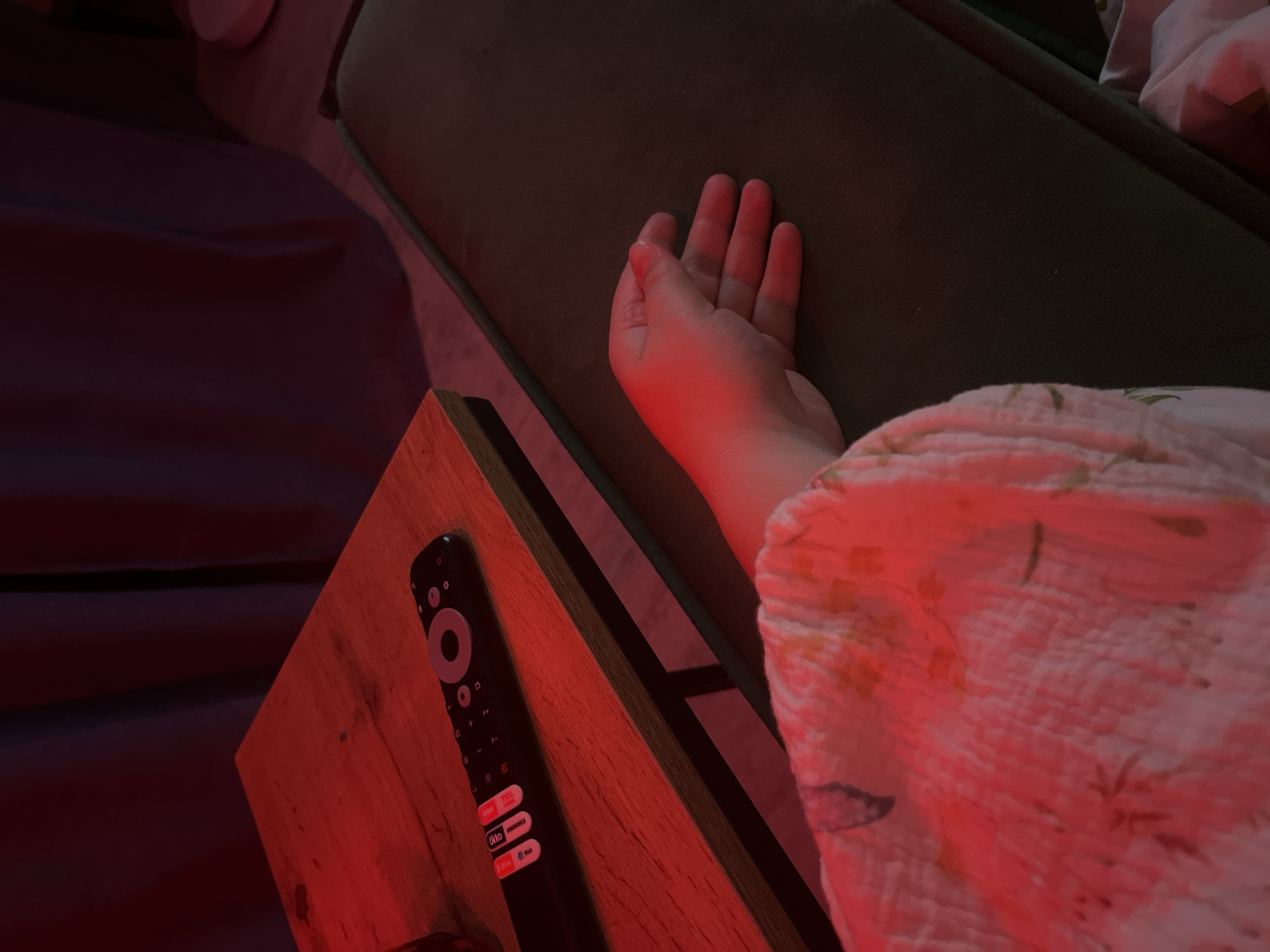 An overhead shot captures a cozy, dimly lit indoor scene, likely a living room or bedroom in Chaikovsky, Russia, bathed in a dominant red and orange ambient light. The low light suggests it is nighttime or late evening.
In the foreground, a person's relaxed right hand and lower forearm are visible, illuminated by the red light, resting on a dark green upholstered surface, possibly a sofa or bed. To the right of the hand, a light-colored, soft blanket or quilt with a subtle floral or leaf pattern is draped, also tinted red by the pervasive lighting.
A wooden side table is positioned to the left of the person's arm. On this table lies a black remote control, prominently displaying a large grey circular button and several app shortcut buttons. Clearly visible text on these buttons includes "TCL," "Disney+", "Rakuten TV," "YouTube," and "Netflix." The red light strongly reflects off the wooden surface and the remote. Further to the left, a dark purple or red sheet or blanket is partially visible, contributing to the room's warm, subdued palette.
The scene conveys a sense of quiet relaxation and leisure, suggesting the person is resting or enjoying entertainment in a comfortable, dimly lit space.