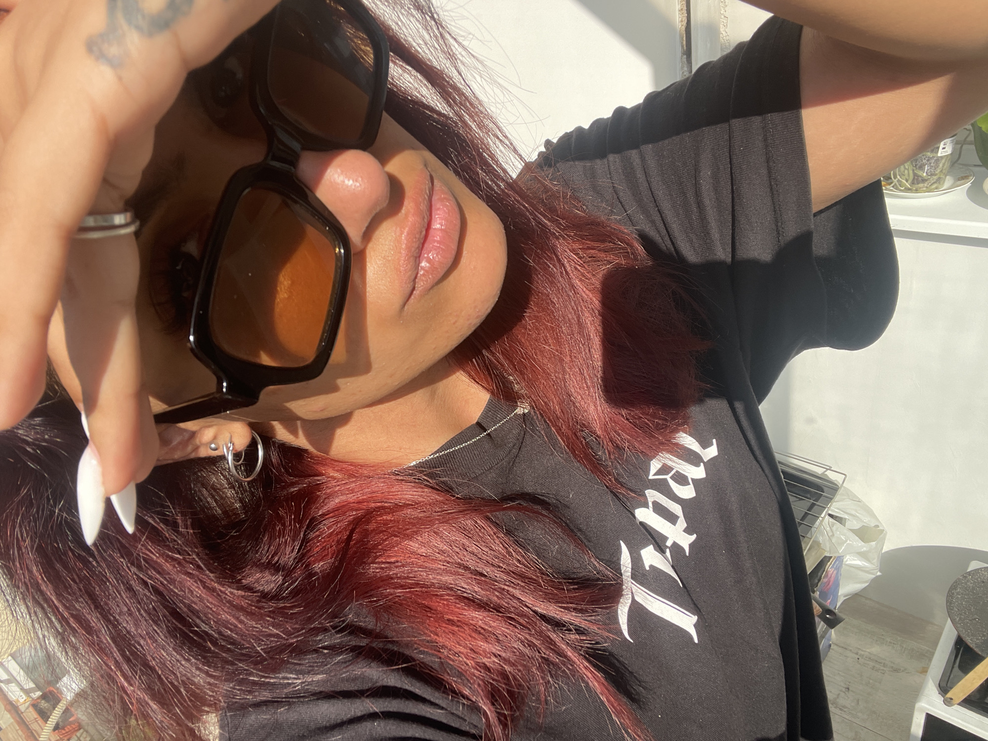 A selfie photograph depicts a person with auburn hair, wearing sunglasses with dark frames and amber lenses, and a black t-shirt. The person's right hand is raised towards the camera, obscuring part of their face. They are adorned with rings on their fingers and multiple piercings on the ear closer to the camera.
Behind the person, a white wall is visible, partially illuminated by sunlight. The scene is indoors, with a glimpse of a stove and other household items visible in the background, suggesting a kitchen setting. The specific location within the city of Unknown, Unknown cannot be determined from the image.