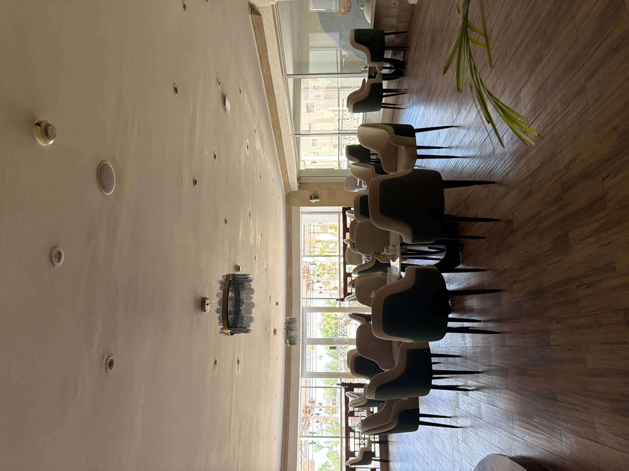 The image depicts the interior of a contemporary lounge area, likely a hotel lobby or restaurant, situated in Sharm El Sheikh, Egypt. The scene is characterized by a warm, neutral color palette dominated by beige, cream, and dark green hues.
The foreground features a polished wooden floor laid in a herringbone pattern. Several plush armchairs, upholstered in a combination of light beige and dark green fabric, are arranged in small clusters around low-lying tables. One table, visible in the center of the frame, is made of white marble with a dark base and holds a small potted plant. Another table, to the right, is a darker, more angular design.
The midground is dominated by a large glass-paneled wall that looks out onto an outdoor area with greenery and architectural structures, suggesting a resort or hotel setting. Two identical modern chandeliers with a circular design and glass accents hang from the ceiling.
The ceiling is white and features several recessed lights and what appear to be smoke detectors. The overall ambiance is one of relaxed sophistication, with ample natural light flooding in from the large windows. There are no people visible in the image.