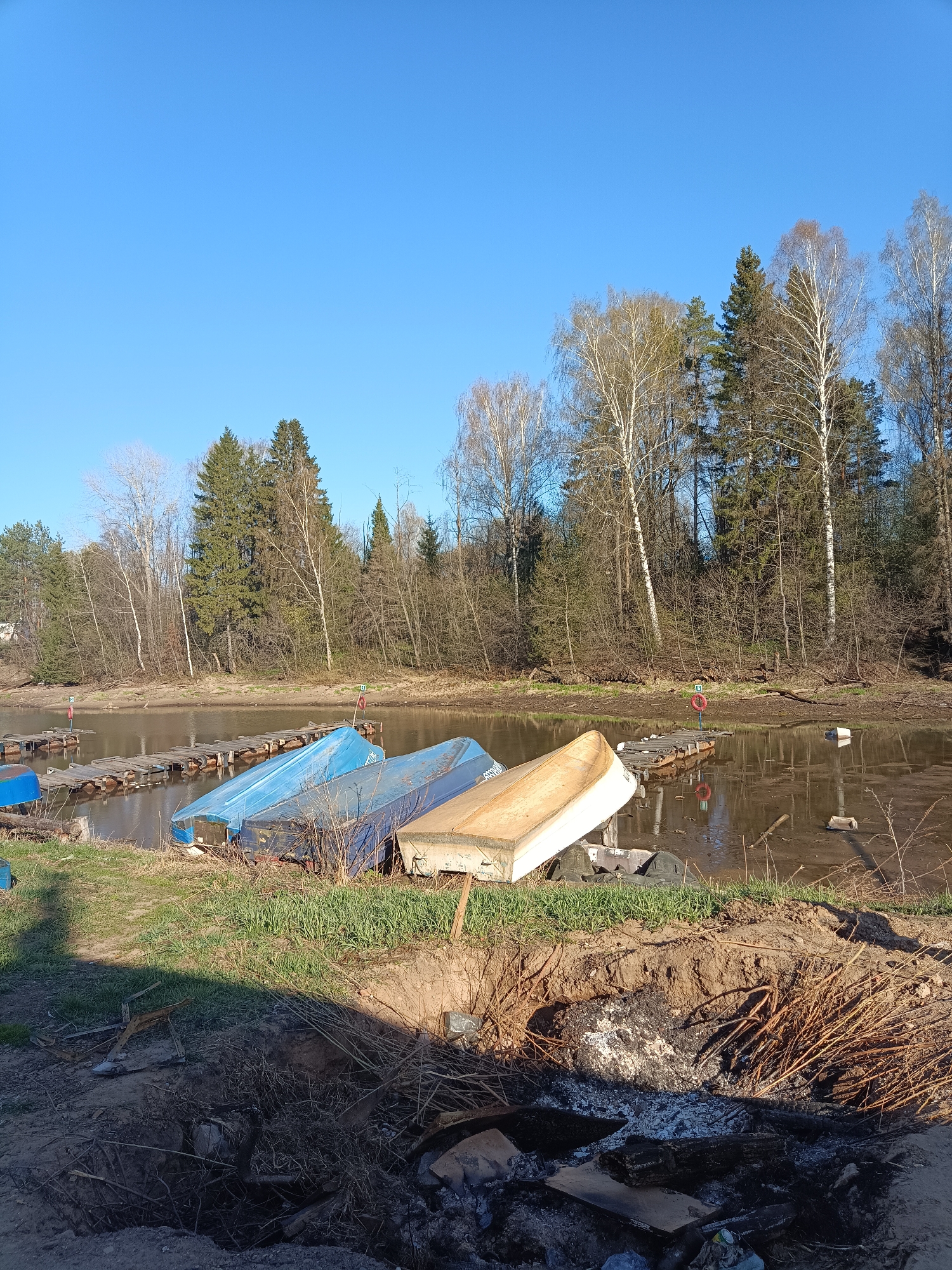 The image shows a waterfront scene in Chaikovsky, Russia. In the foreground, there is a disturbed area, possibly a former campfire site, with charred wood, ash, and discarded trash. Behind this, three overturned boats rest on the grassy bank of a body of water. Two boats are blue and one is off-white. The water is murky brown and appears to be at a low level, exposing wooden docks or platforms supported by pilings. Beyond the water, a mixed forest of deciduous and evergreen trees lines the opposite bank. The sky is clear and blue. There are no people visible in the image. The scene suggests a recreational area, possibly a boat launch or small harbor that may have experienced recent use and neglect.