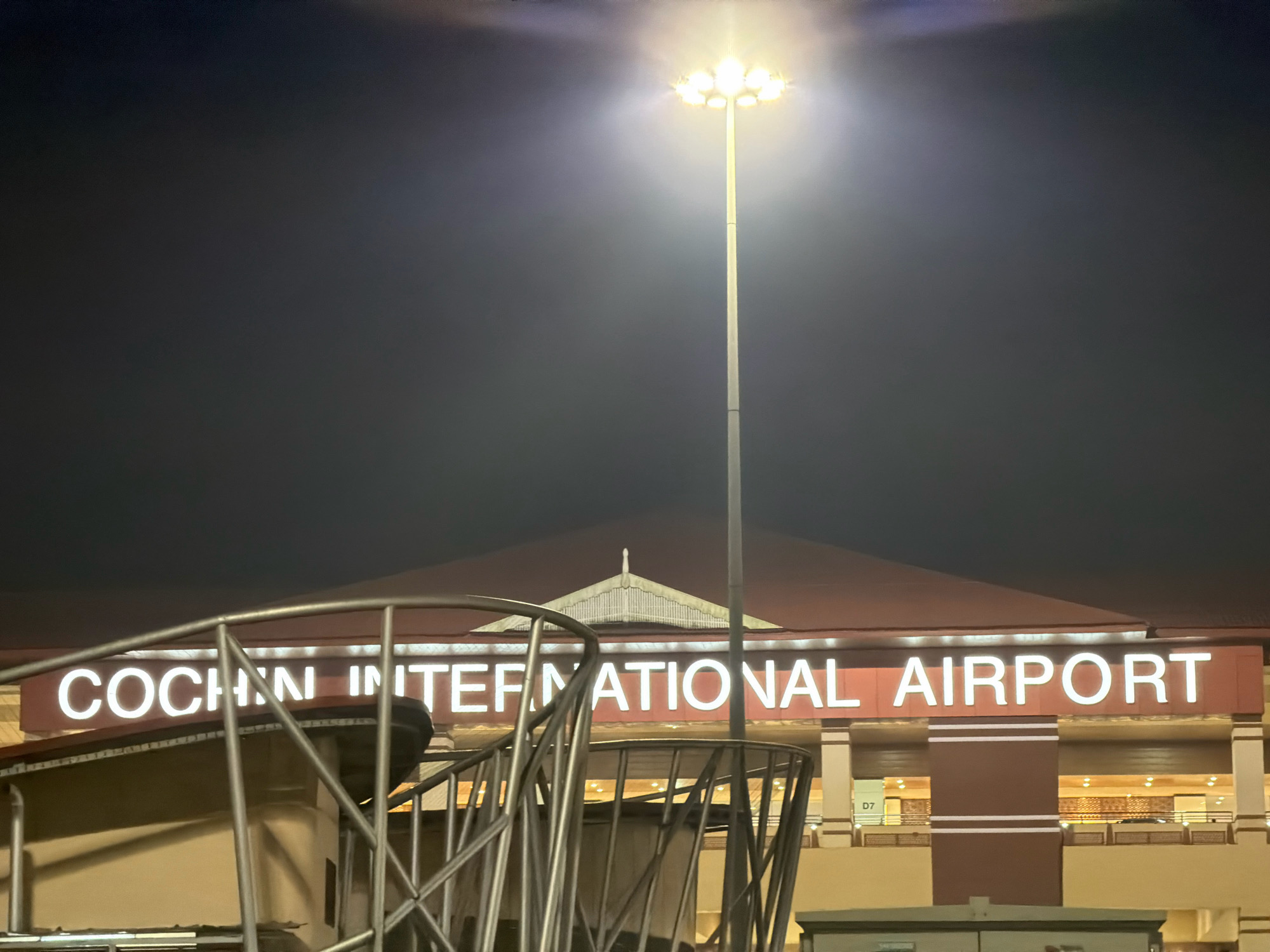 The image shows the Cochin International Airport in Kochi, India at night. A tall, modern light pole with multiple bright lights illuminates the area, casting a bright glow upwards and outwards into the dark sky. Below the lights, the facade of the airport building is visible, with a prominent sign reading "COCHIN INTERNATIONAL AIRPORT" in large, illuminated white letters against a dark red background. The airport structure features a distinctive roofline and architectural details, with striped brown and beige sections. In the foreground, a section of a metallic railing is visible, suggesting the vantage point is from a higher level or a vehicle. The overall scene captures the illuminated entrance of a major transportation hub at night.
