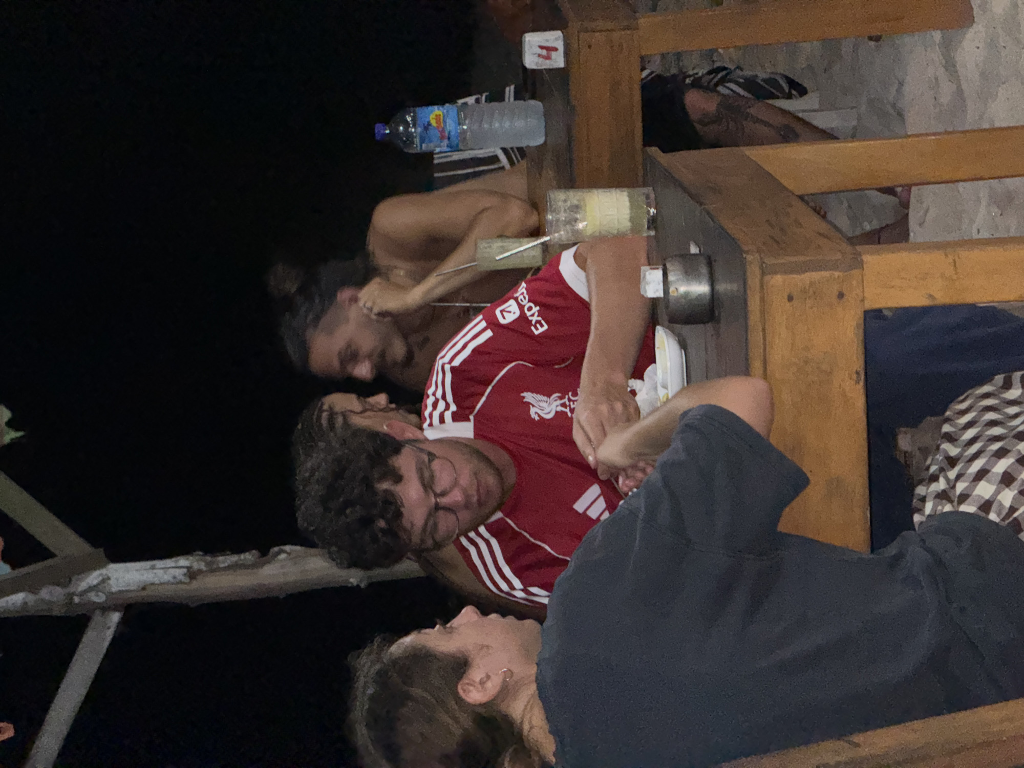 The image shows a group of people at a bar or outdoor seating area in Unknown, Indonesia. Three individuals are clearly visible. One man wears a red Liverpool soccer jersey with white stripes. He is wearing glasses and has curly hair. Another man with dark hair is shirtless and appears to be laughing or speaking. A woman is sitting and wearing a dark t-shirt. A wooden table is visible with items on it, including a water bottle and glassware. The setting is dark, suggesting it's nighttime. The environment appears to be a casual outdoor space with simple wooden structures.