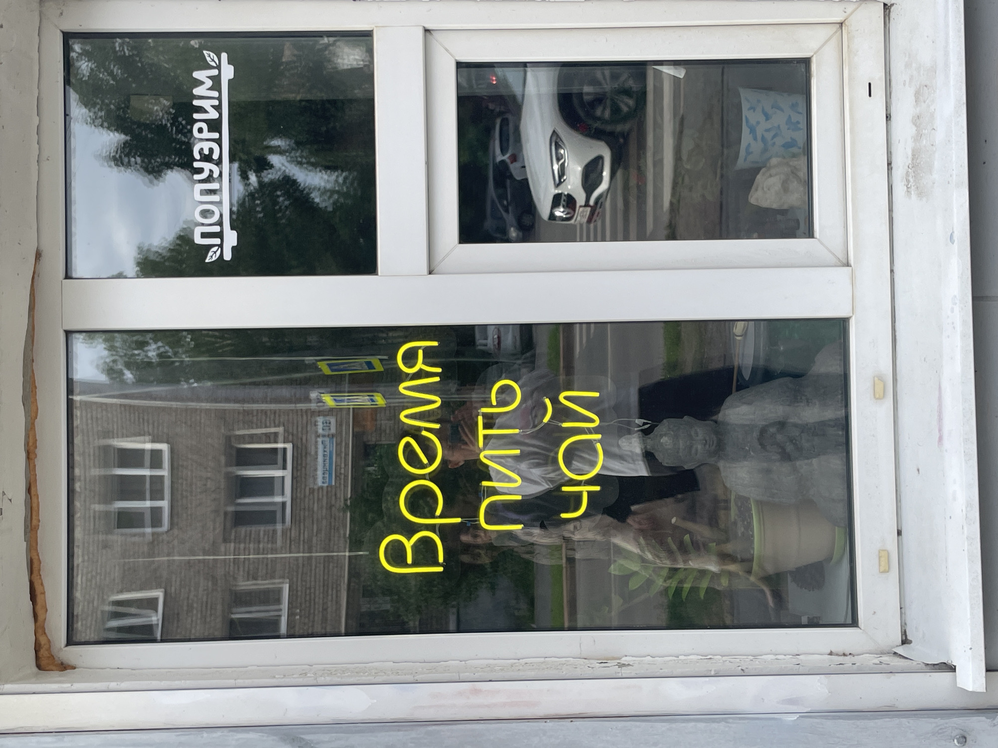 In Izhevsk, Russia, a storefront window displays a neon yellow sign in Cyrillic script that reads "Время пить чай" which translates to "Time to drink tea." To the right of this sign, on a separate pane of glass, is a white graphic featuring the Cyrillic word "ПОПУЗРИМ" with decorative leaves on either side. Reflected in the window is a building across the street, trees, and passing vehicles. Inside the storefront, visible through the window, are a Buddha statue, a potted plant, and a person taking a photograph with their phone. The window frame appears weathered and shows signs of age.