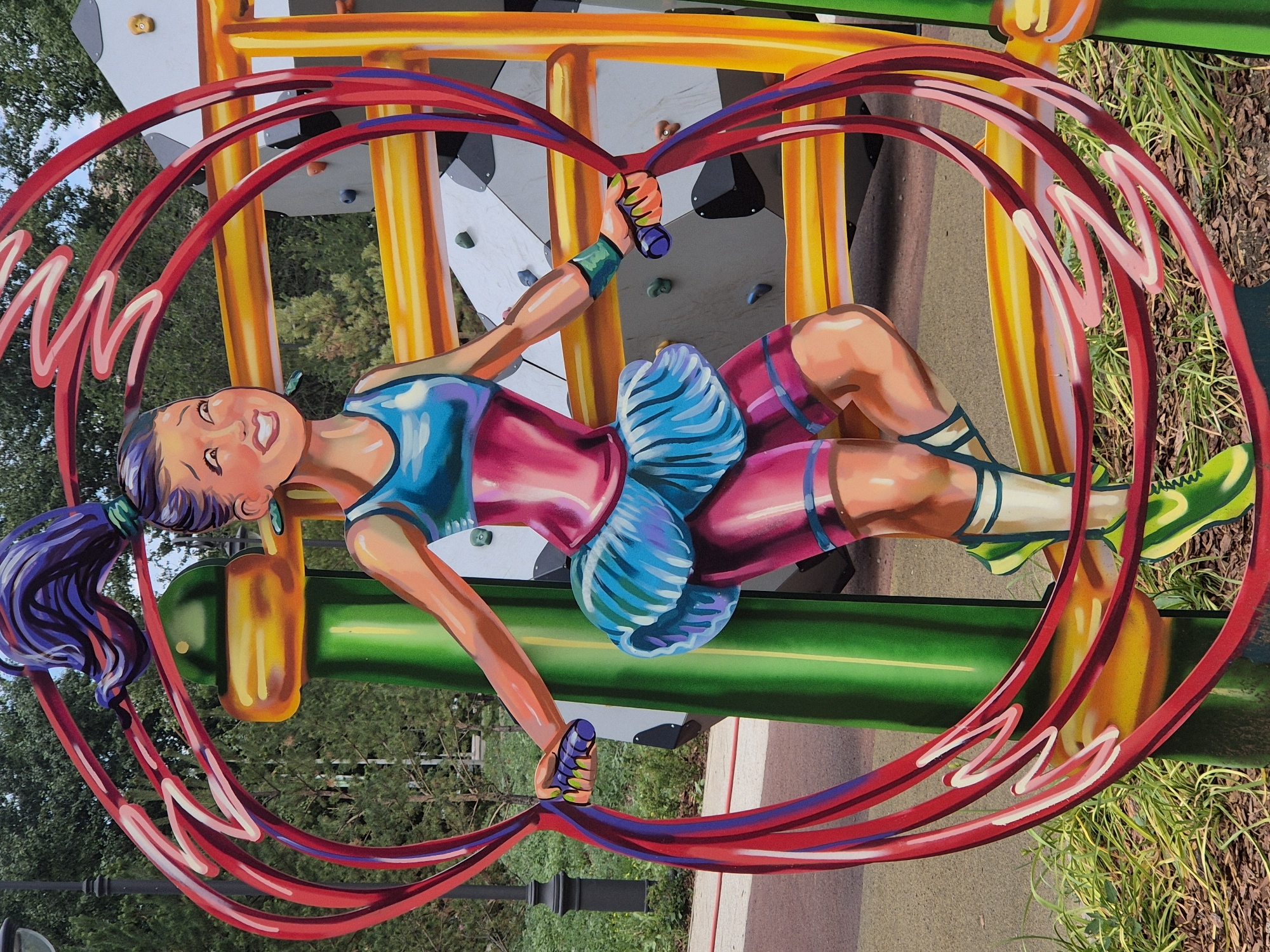 This is a photograph of a colorful mural depicting a cartoonish girl jumping rope. The girl has dark purple hair styled in a high ponytail, a wide smile, and is wearing a blue and pink athletic outfit with a blue ruffled skirt. She is surrounded by a stylized red jump rope that forms a large loop. The mural is placed outdoors, likely as part of a playground, with elements of playground equipment visible in the background, including yellow bars and a grey climbing wall with colorful handholds. The environment includes trees, grass, and a paved walkway. The mural is brightly painted, with a strong emphasis on shading and highlights to give the figure a three-dimensional appearance. The location of this mural is in the city of Kokshetau, Kazakhstan, as indicated by the metadata.
