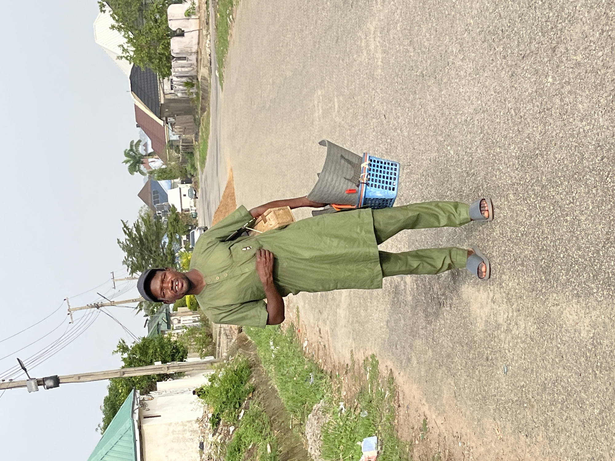 A man stands on a paved road in Abuja, Nigeria. He is wearing an olive green tunic and trousers, a dark cap, and grey sandals. He is carrying a blue and grey crate containing miscellaneous items. The road is lined with green vegetation and buildings. Power lines are visible overhead.
