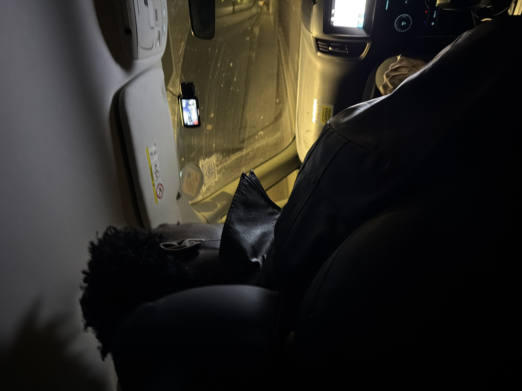 A person with dark, curly hair is seated in the passenger seat of a vehicle at night. They are wearing a black leather jacket. The vehicle's interior is visible, including the dashboard, windshield, and a digital display screen. A dashcam is mounted on the windshield. The scene appears to be in a city, possibly London, United Kingdom, given the nighttime lighting and urban environment suggested by the blurry background.