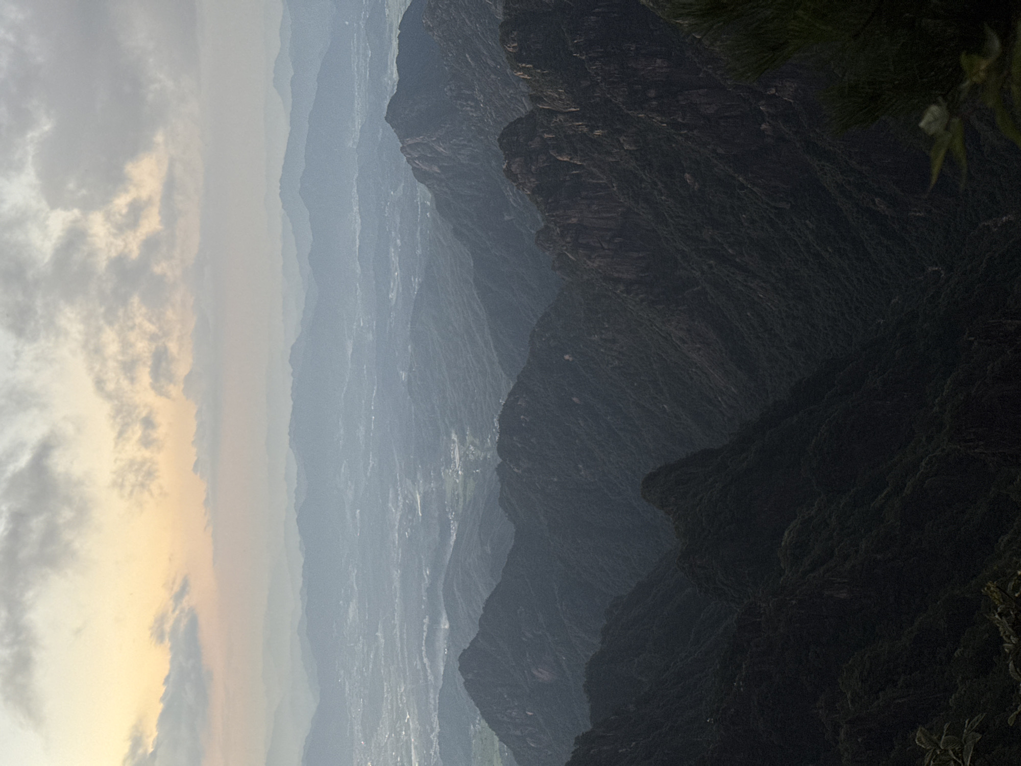 This is a high-angle landscape photograph of layered mountains under a cloudy sky at dusk. The foreground features dark, densely forested mountain slopes with rugged rock formations visible. The middle ground shows more distant, less distinct mountain ranges fading into a hazy, pale sky tinged with yellow and pink, suggesting either sunrise or sunset. In the far distance, a valley or plain is visible with scattered lights indicating a settlement, likely the city of Huangshan District, China. The overall atmosphere is serene and vast, capturing the expansive natural beauty of the mountainous terrain.