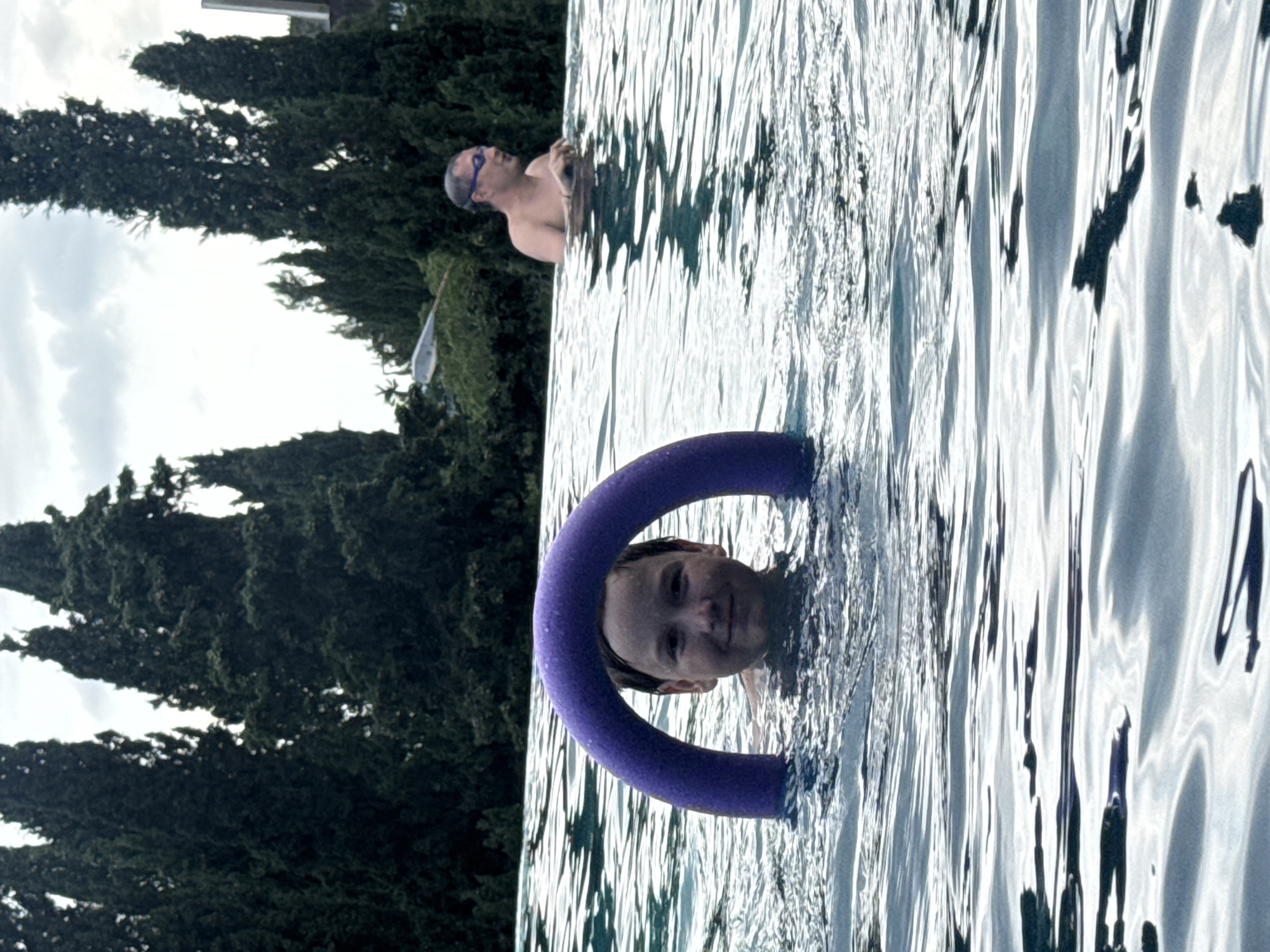 The image shows two individuals in a swimming pool with cypress trees and buildings in the background. In the foreground, a young boy is partially submerged in the water, with a purple pool noodle forming an arc around his head. He is looking directly at the camera with a slight smile. In the background, to the right, a man wearing swimming goggles is also in the pool, looking to his left. The water's surface is rippled, reflecting the overcast sky. The setting appears to be a residential area in the city of Yalta, Ukraine, given the characteristic cypress trees and architecture often found in the region.