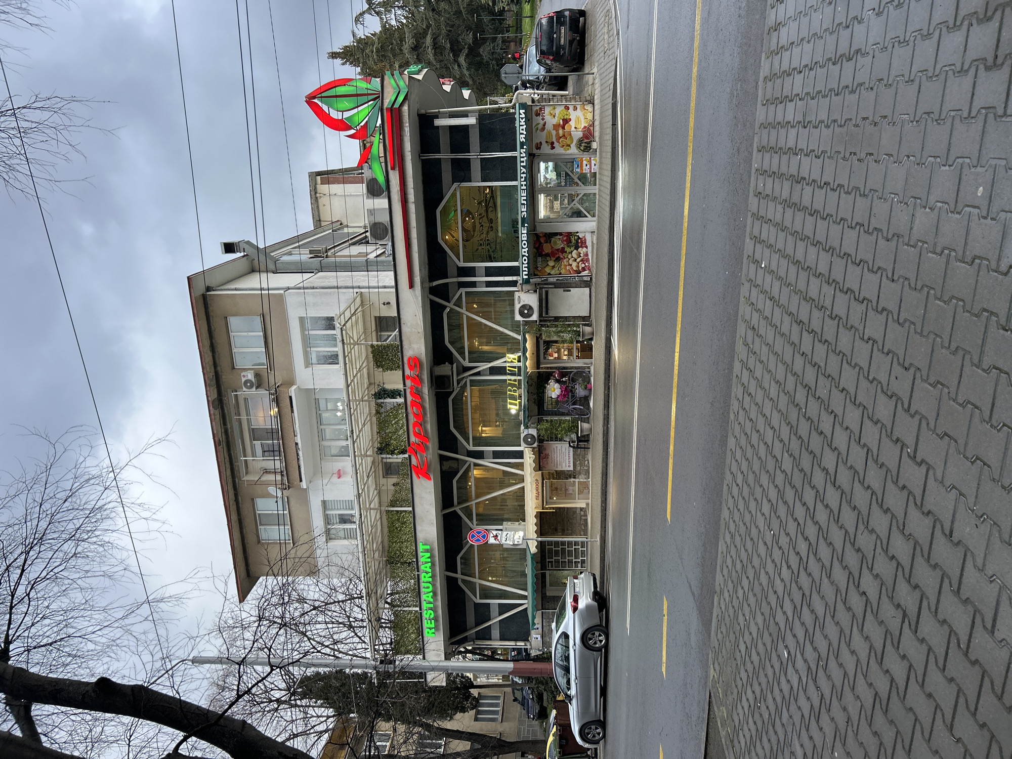 A street scene in Stara Zagora, Bulgaria, features a multi-story building housing a restaurant called "Ripariso." The restaurant is distinguished by red and green signage, and a large fruit and vegetable display on the ground floor. A silver car is parked on the street in front of the restaurant. The street is marked with yellow and white lines. A paved sidewalk is visible on the right. Trees and power lines are also present. The sky is overcast.