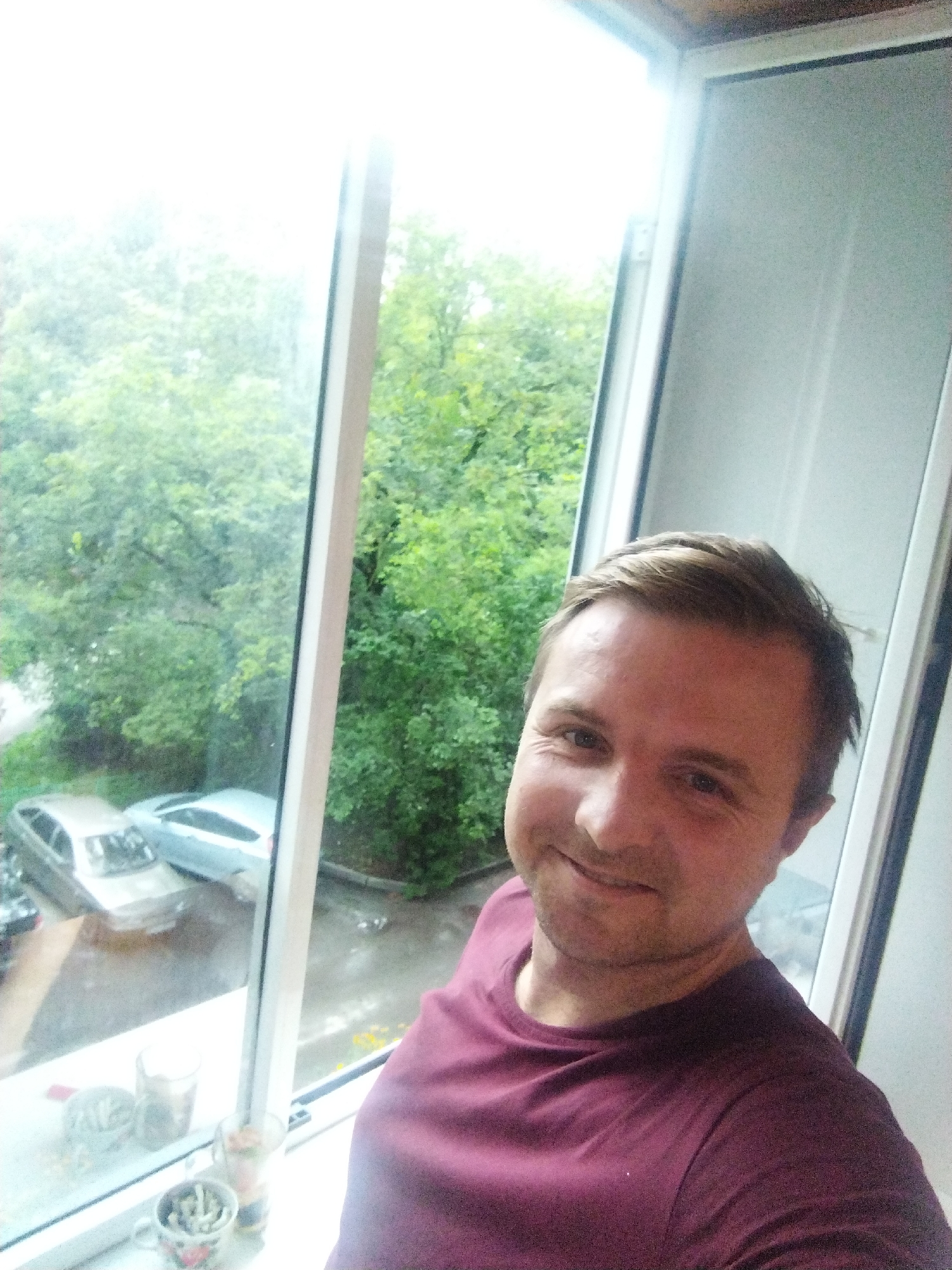 A man is taking a selfie from inside a building, looking out of a window. He is positioned in the foreground of the image, with his face and upper body visible. The window reveals a view of trees and parked cars outside, suggesting an urban or suburban setting. The lighting is bright, possibly indicating daylight. The man is wearing a maroon t-shirt. The scene appears to be located in Lipetsk, Russia.