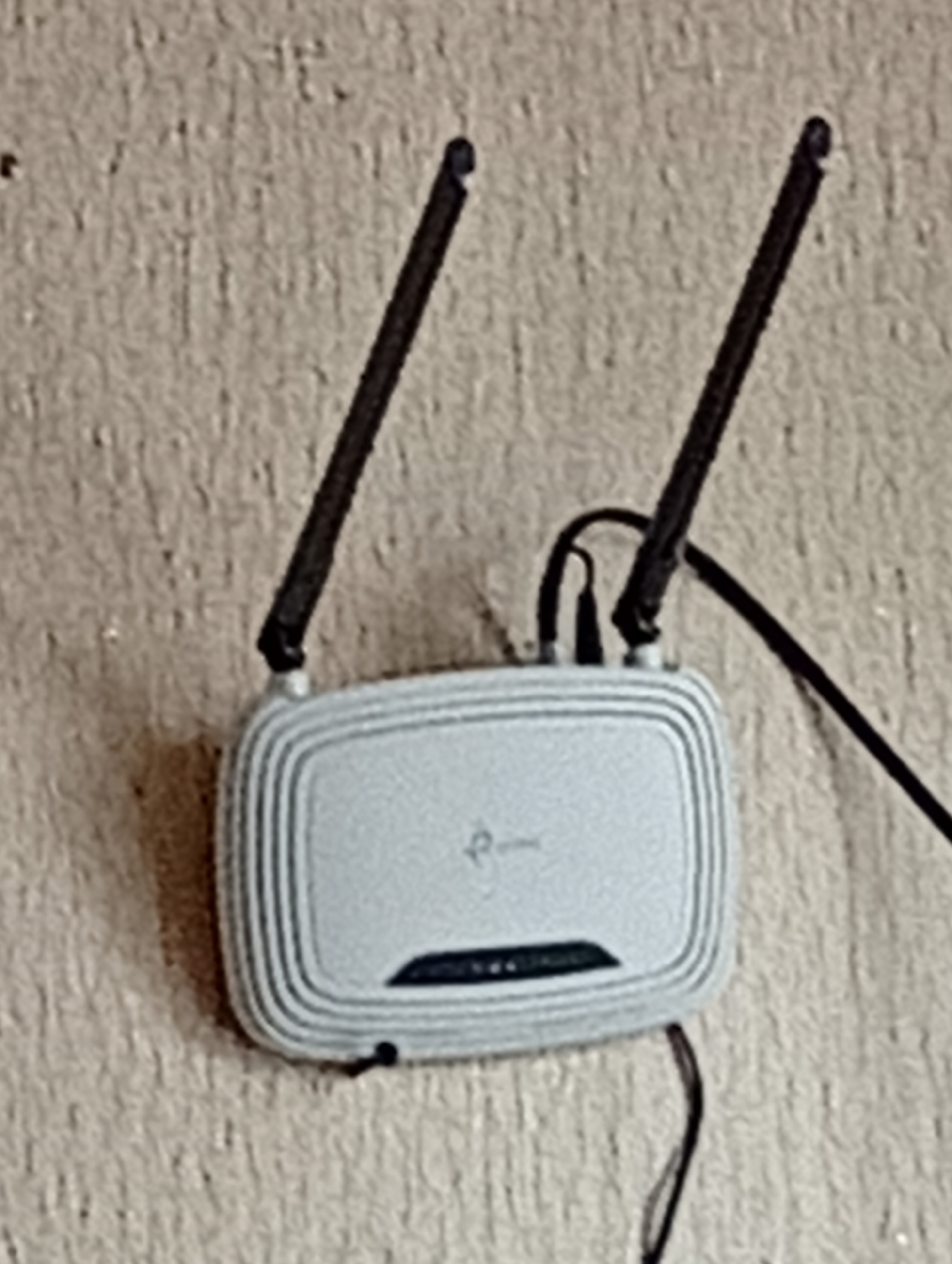 A close-up, blurry, and overexposed image shows a silver TP-Link Wi-Fi router mounted on a textured, light-colored wall. The router has two black antennas extending upwards. Cables are connected to the router, with one black cable extending downwards and to the right. The lighting is harsh, creating a washed-out appearance and obscuring finer details of the wall texture and the router's features. The image appears to be taken indoors.